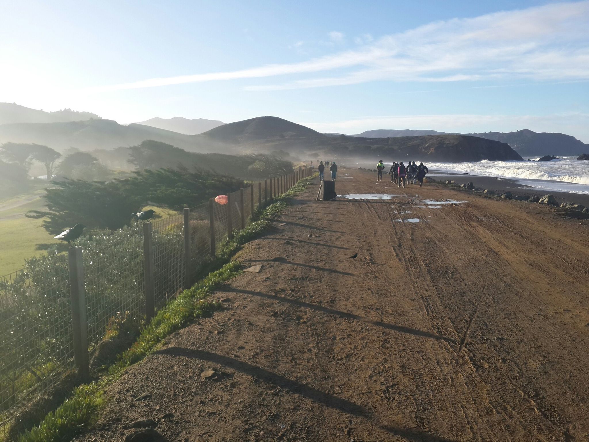 Group of people walking on a wide dirt road beside a misty ocean. - Mori Point Headlands – Sharp Park Beach loop from Milagra Ridge