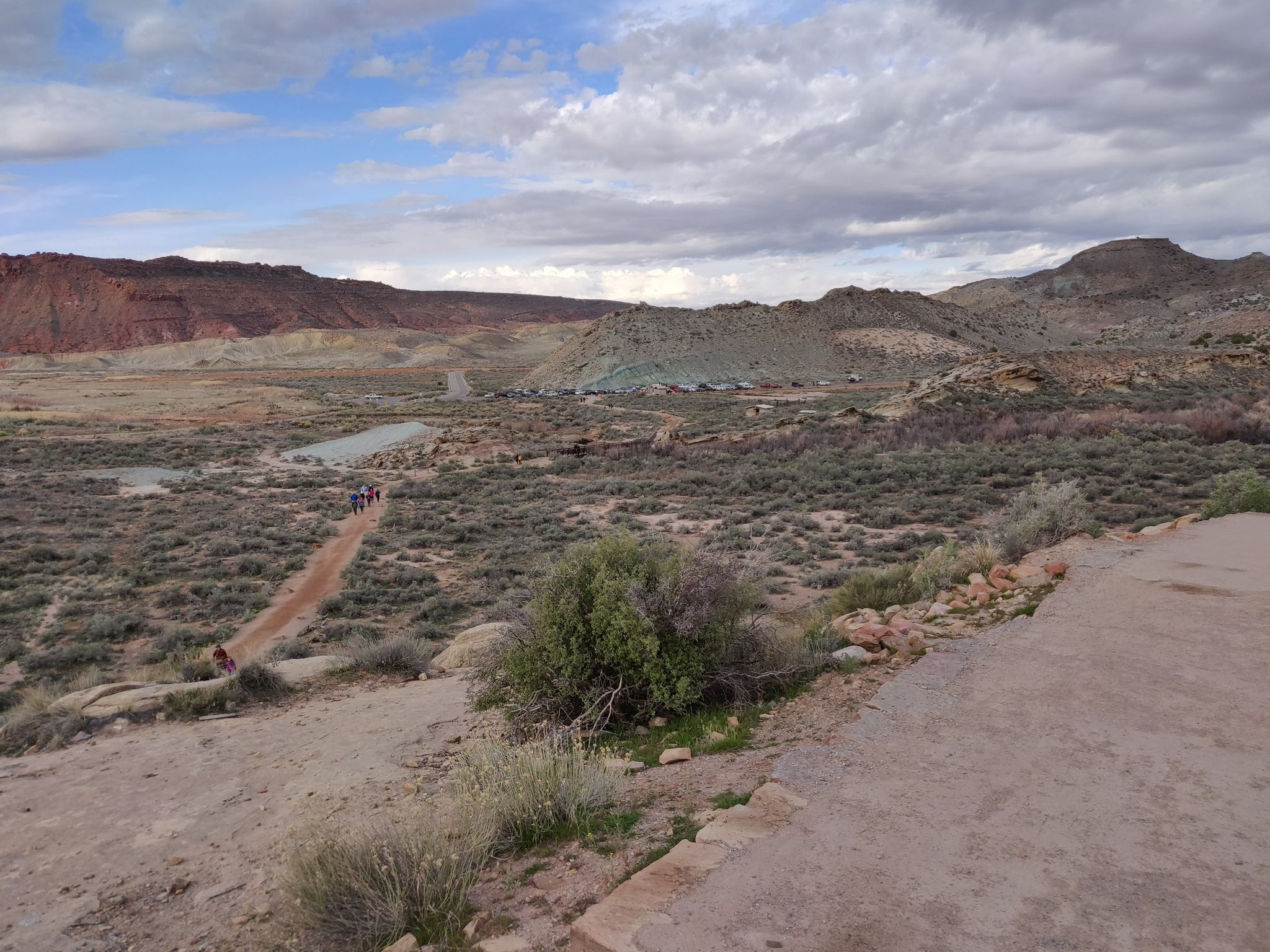 Hikers on a dirt path through a vast desert valley under cloudy skies. - Delicate Arch Trail