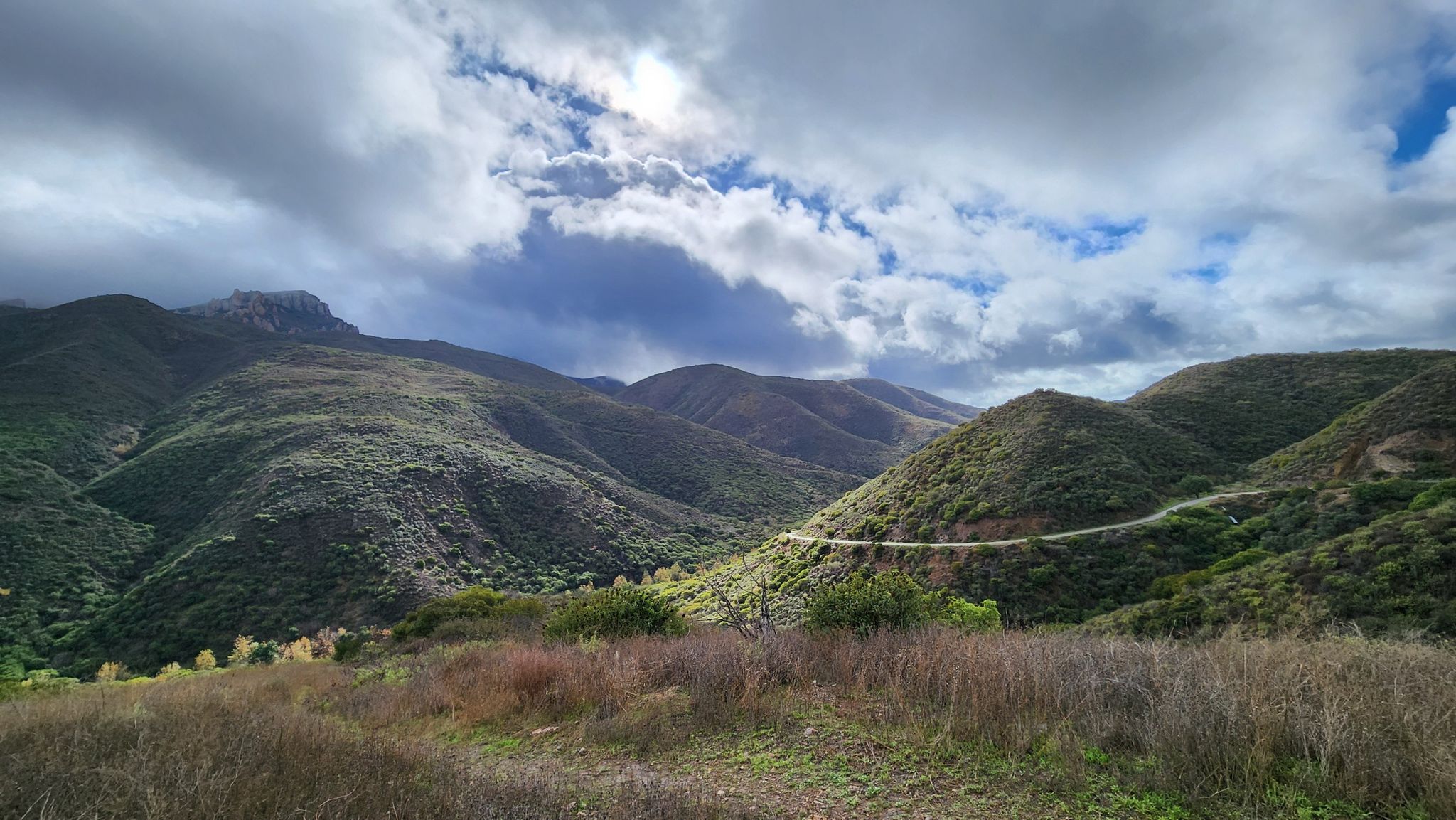 Green mountains under a cloudy sky with a winding road. - Satwiwa Loop and Old Boney Trail