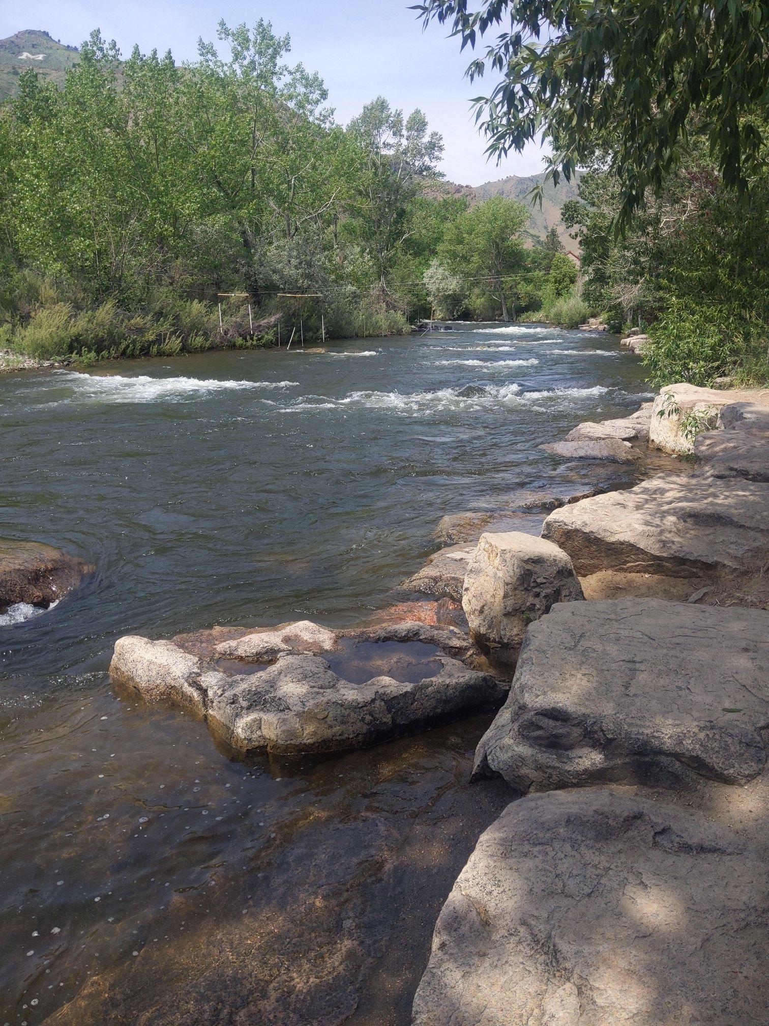 Calm river flowing past large rocks and green trees on a bright day. - Clear Creek Trail loop from Rocky Mountain Quilt Museum
