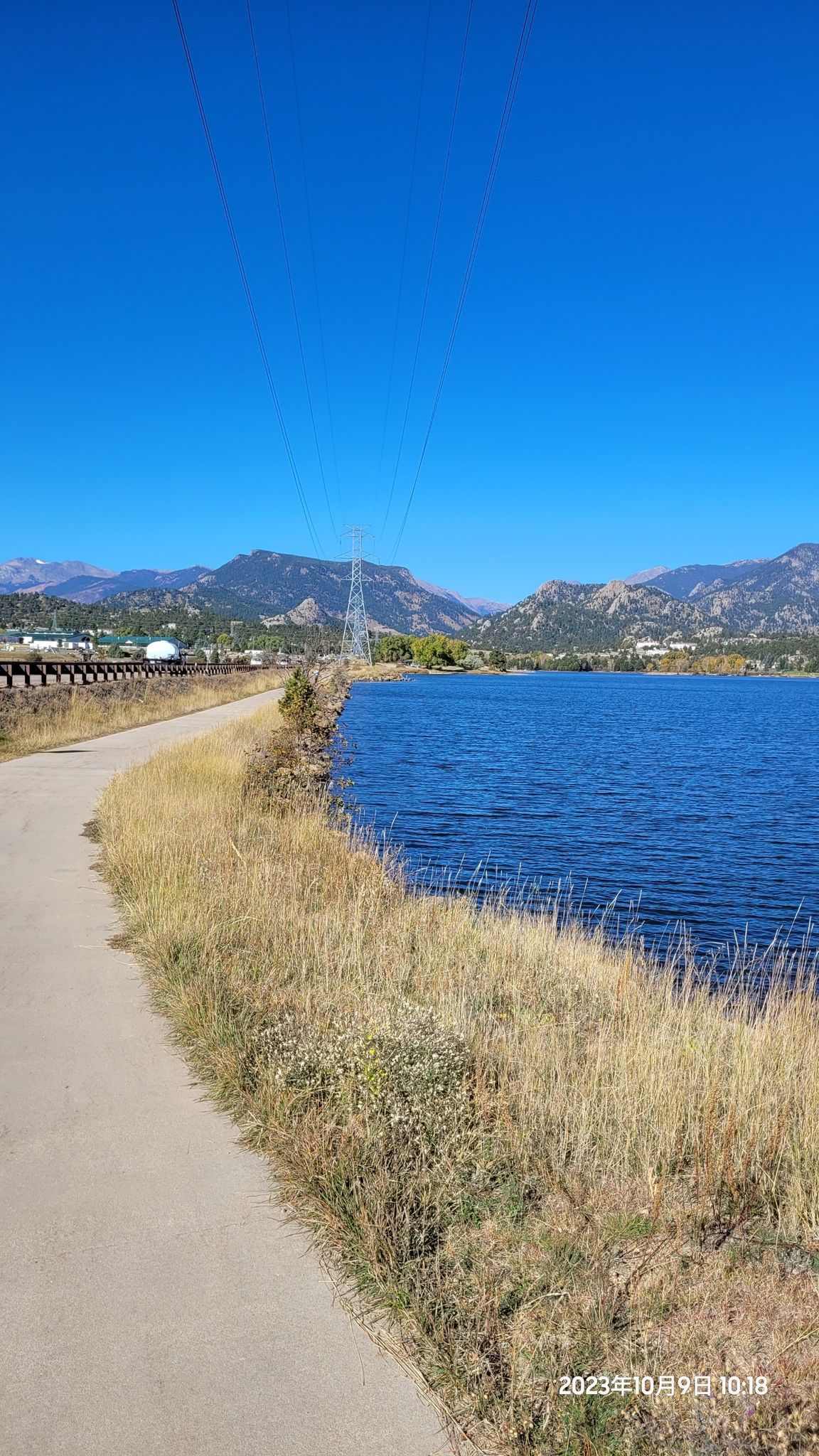 Paved path alongside a lake with mountains and a power tower under blue sky. - Lake Estes Trail