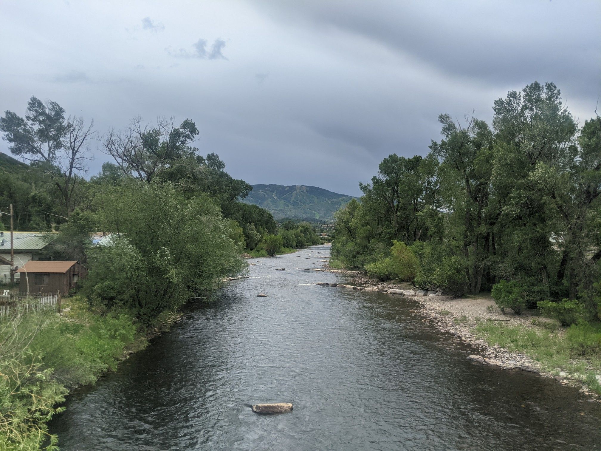 River flows through a wooded area with distant mountains under a dark, cloudy sky. - Yampa River Core Trail