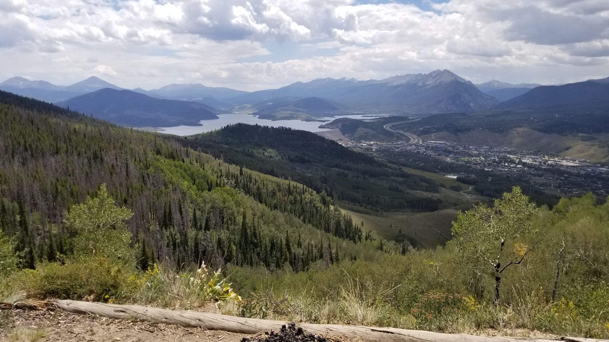 Expansive mountain landscape with a lake, town, and forested slopes under clouds. - Ptarmigan Peak Trail