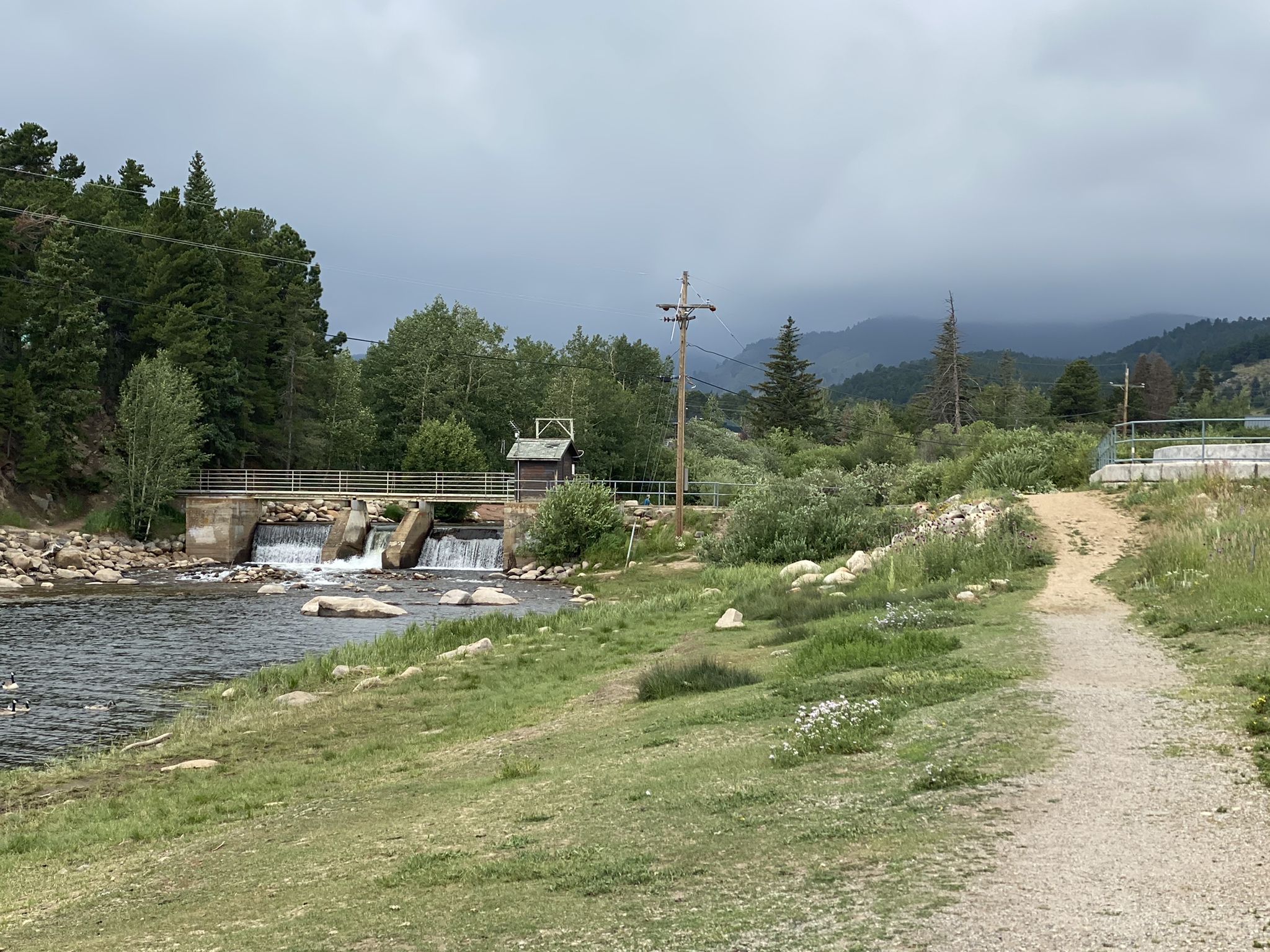 A river flows past a small dam and dirt path under a cloudy sky. - Rollinsville & Barker Reservoir loop from Nederland