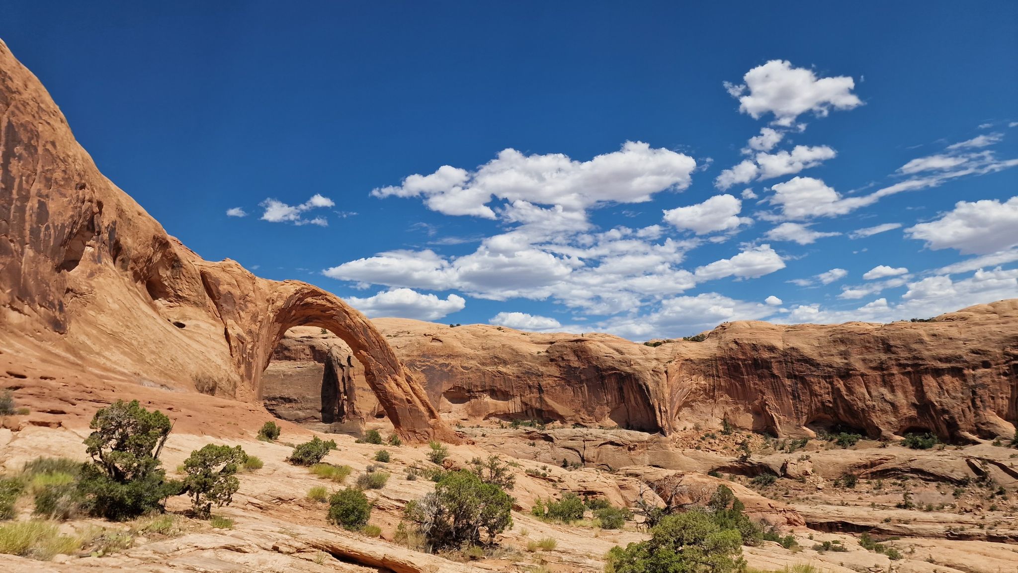 A massive natural rock arch stands in a vast desert landscape under a cloudy sky. - Corona Arch Trail