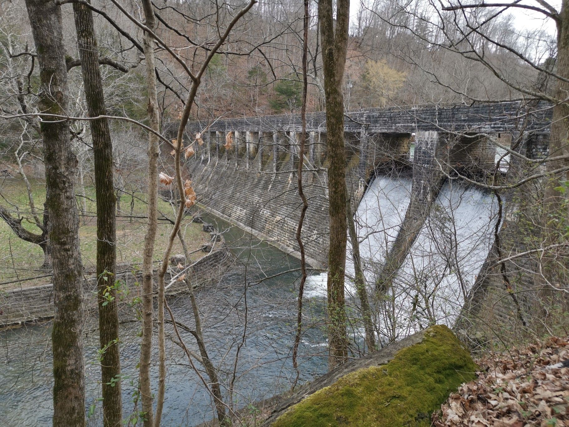 Large stone dam with water flowing over it, viewed through bare trees. - Standing Stone Lake Trail