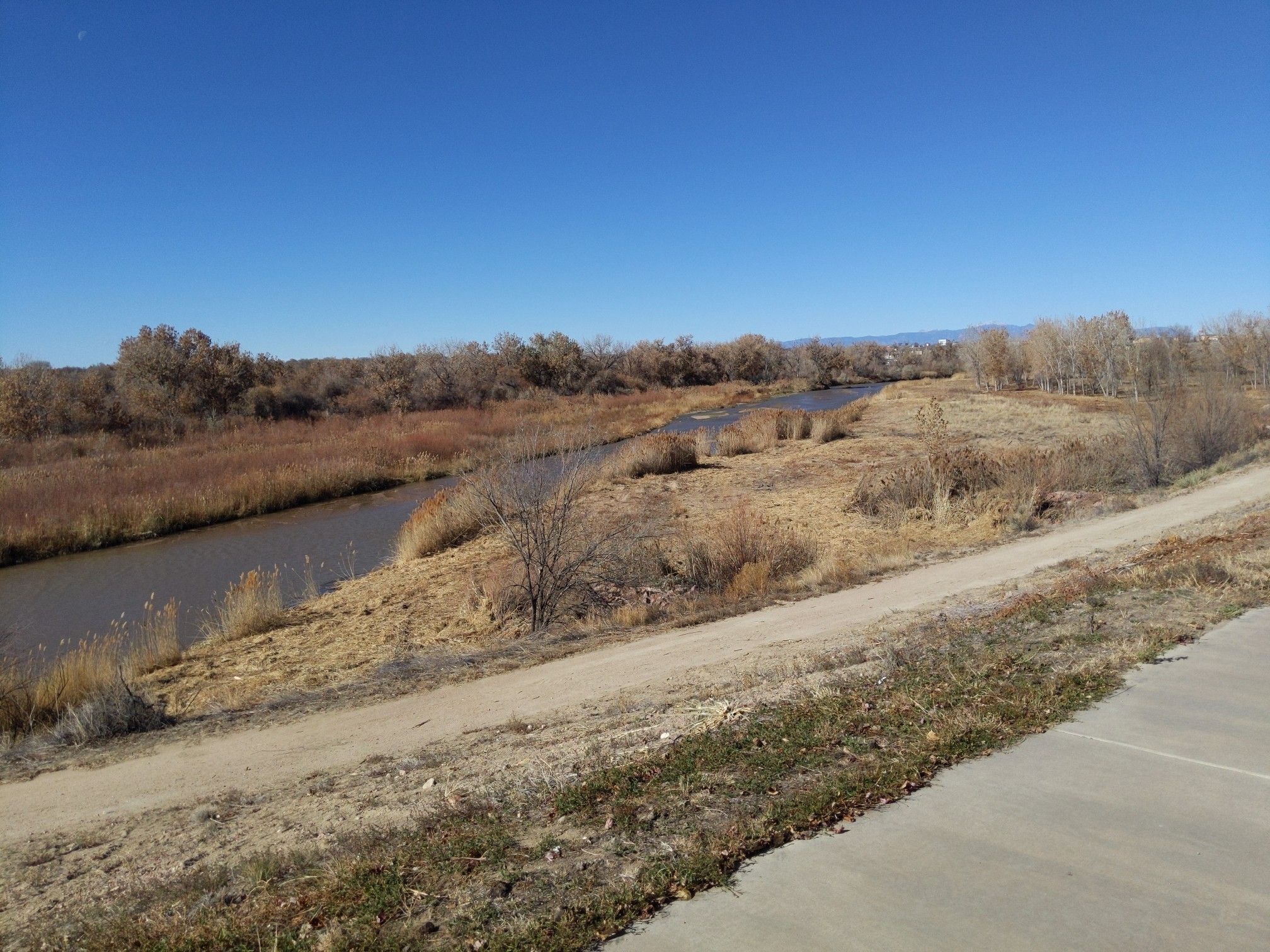 A river flows past dry brown fields and a dirt path under a clear sky. - Fountain Creek Trail