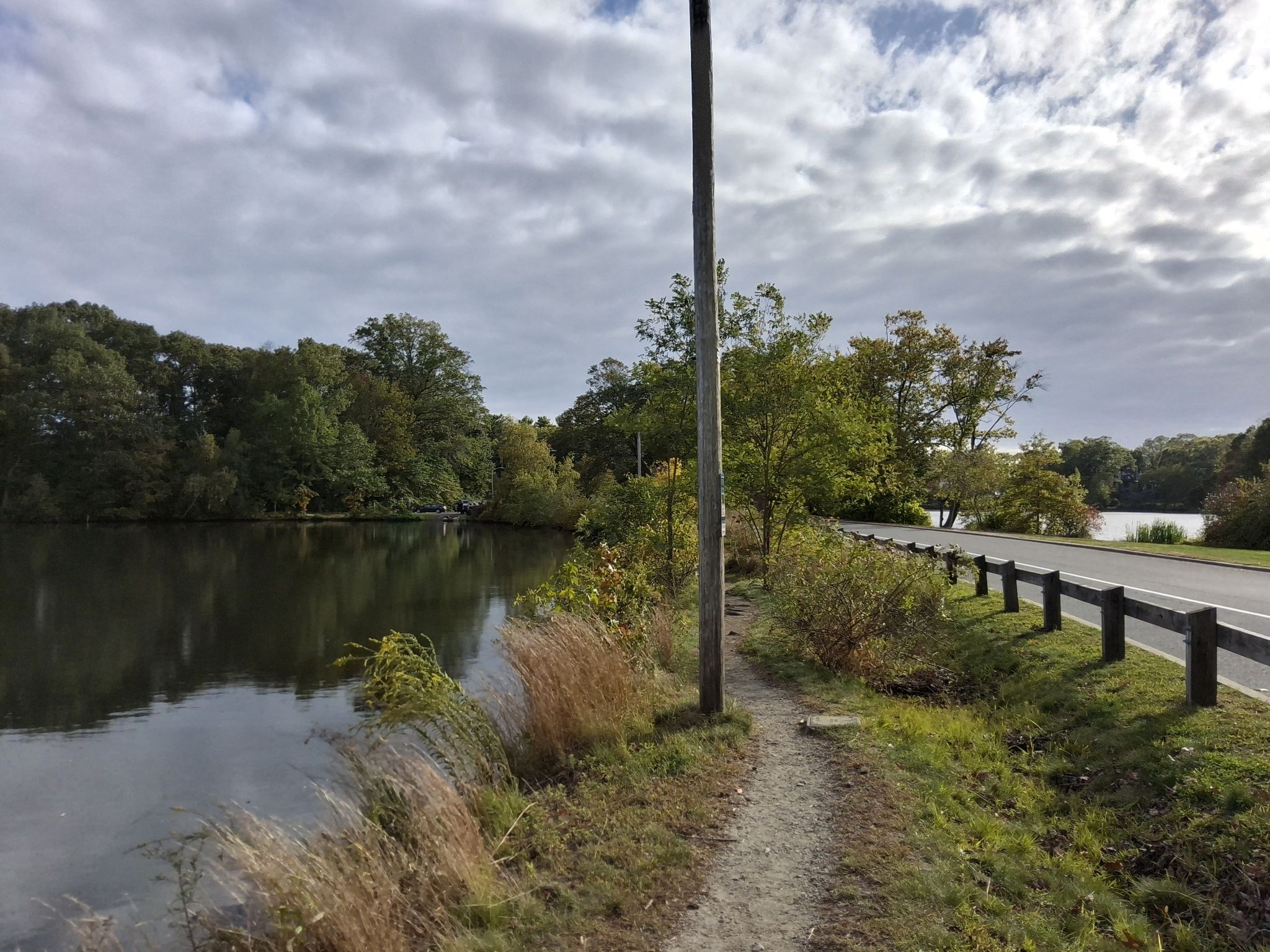 A dirt path runs alongside a lake and road under a cloudy sky. - Roger Williams Park – Elm Lake loop from Johnson and Wales University Providence Harborside Campus