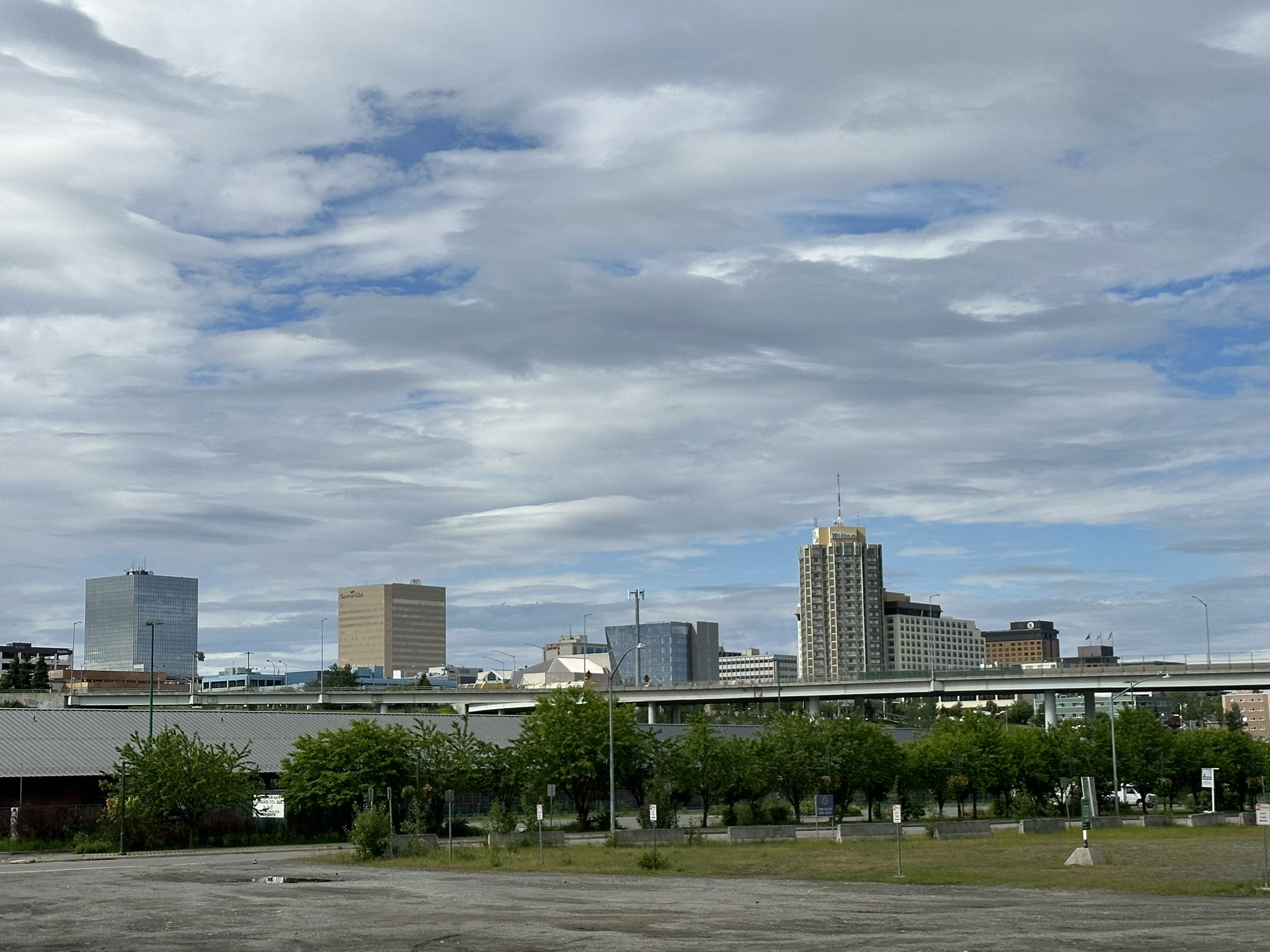 An urban skyline with tall buildings and an overpass under a cloudy sky. - Alaska Railroad locomotive – Ship Creek Trail loop from Anchorage
