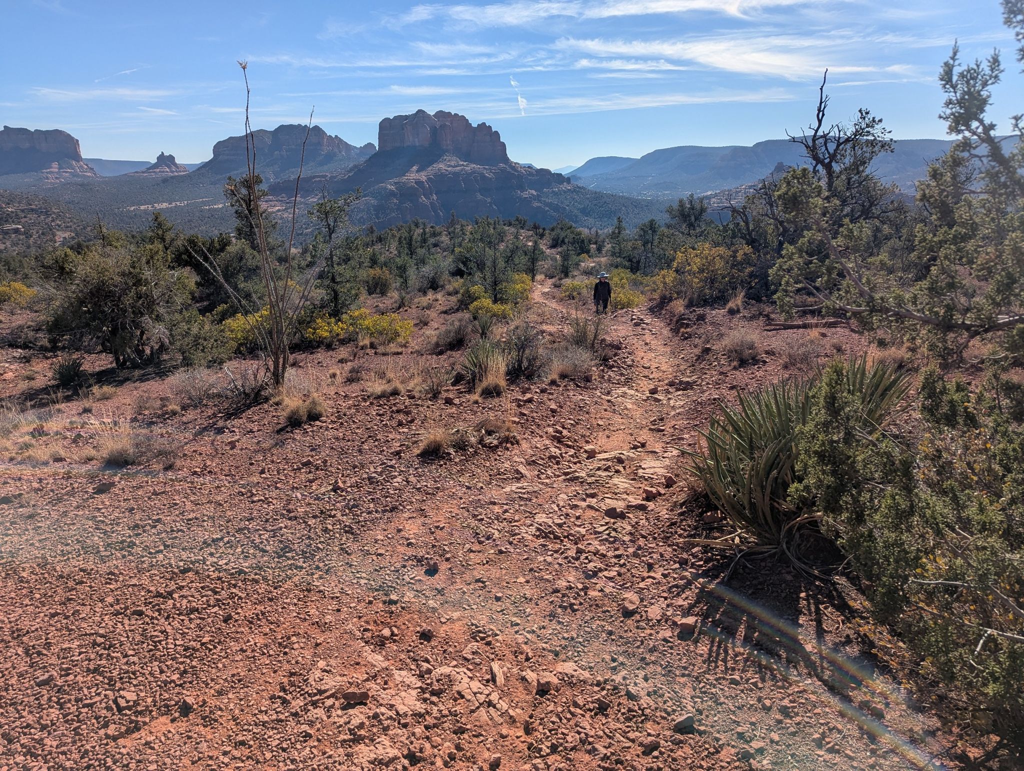 A person walks on a red dirt trail towards distant red rock formations under blue sky. - Ridge Trail (Sedona) loop from Stutz Bearcat Dr (Golden Goose Amer. Grill)