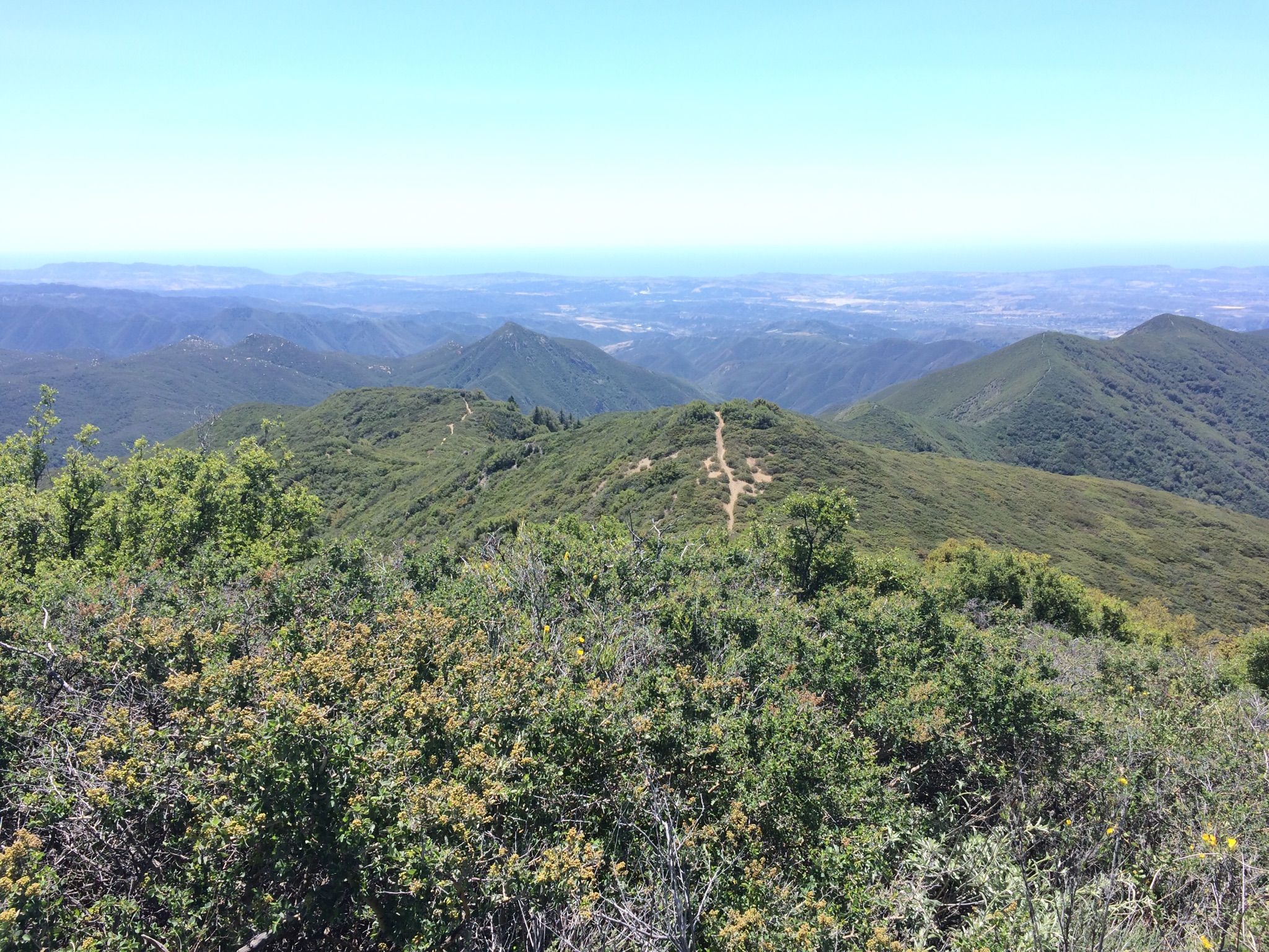 A winding trail across sunlit green mountains with a distant valley under clear skies. - Los Pinos Trail