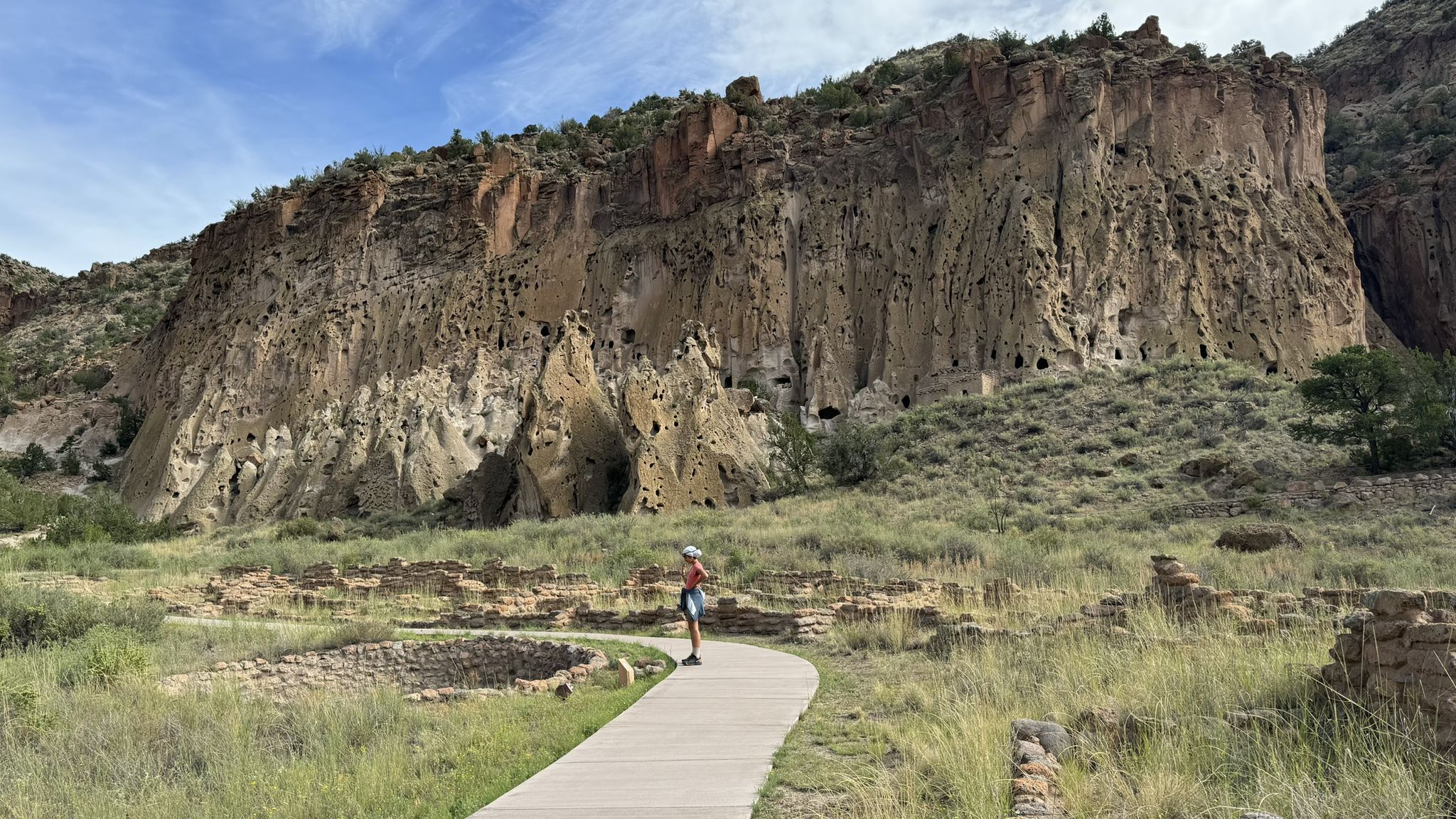 A person stands on a boardwalk facing a massive cliff with many cave dwellings. - Frey Trail