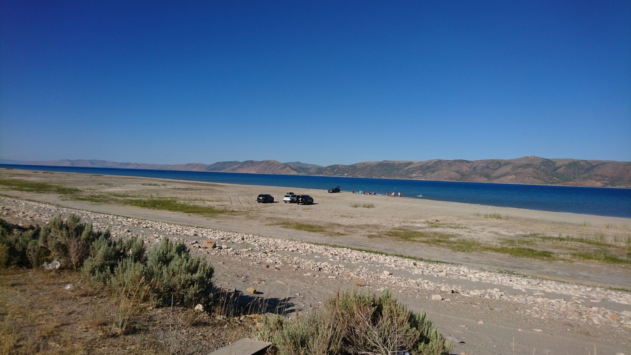 Sunny day, cars and people on a wide, rocky lake shore with distant mountains. - Bear Lake Legacy Pathway