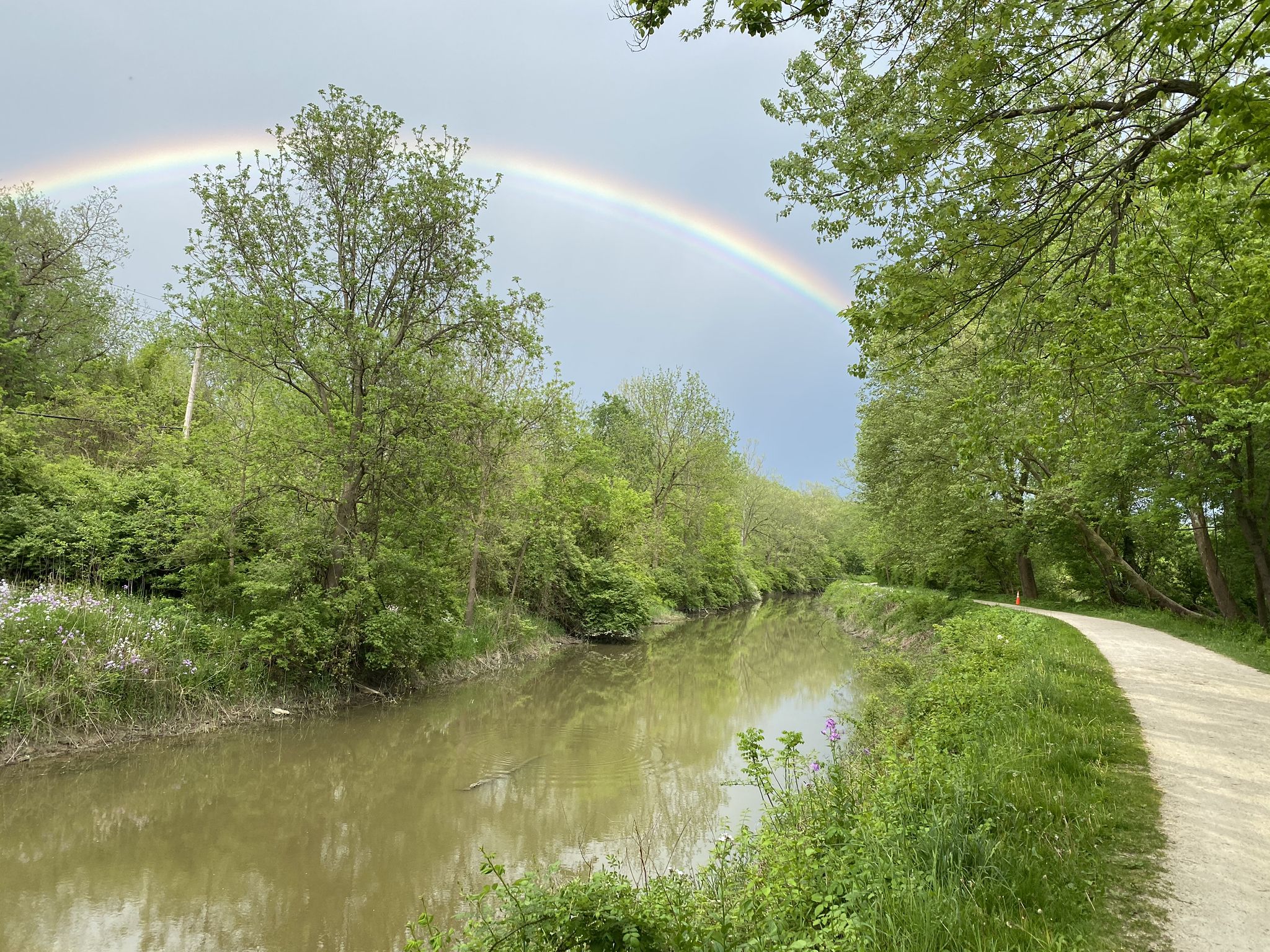 Rainbow over a calm river next to a dirt path, surrounded by green trees. - Lock 39, Ohio & Erie Canal – Bath House Café loop from A Christmas Story House Museum
