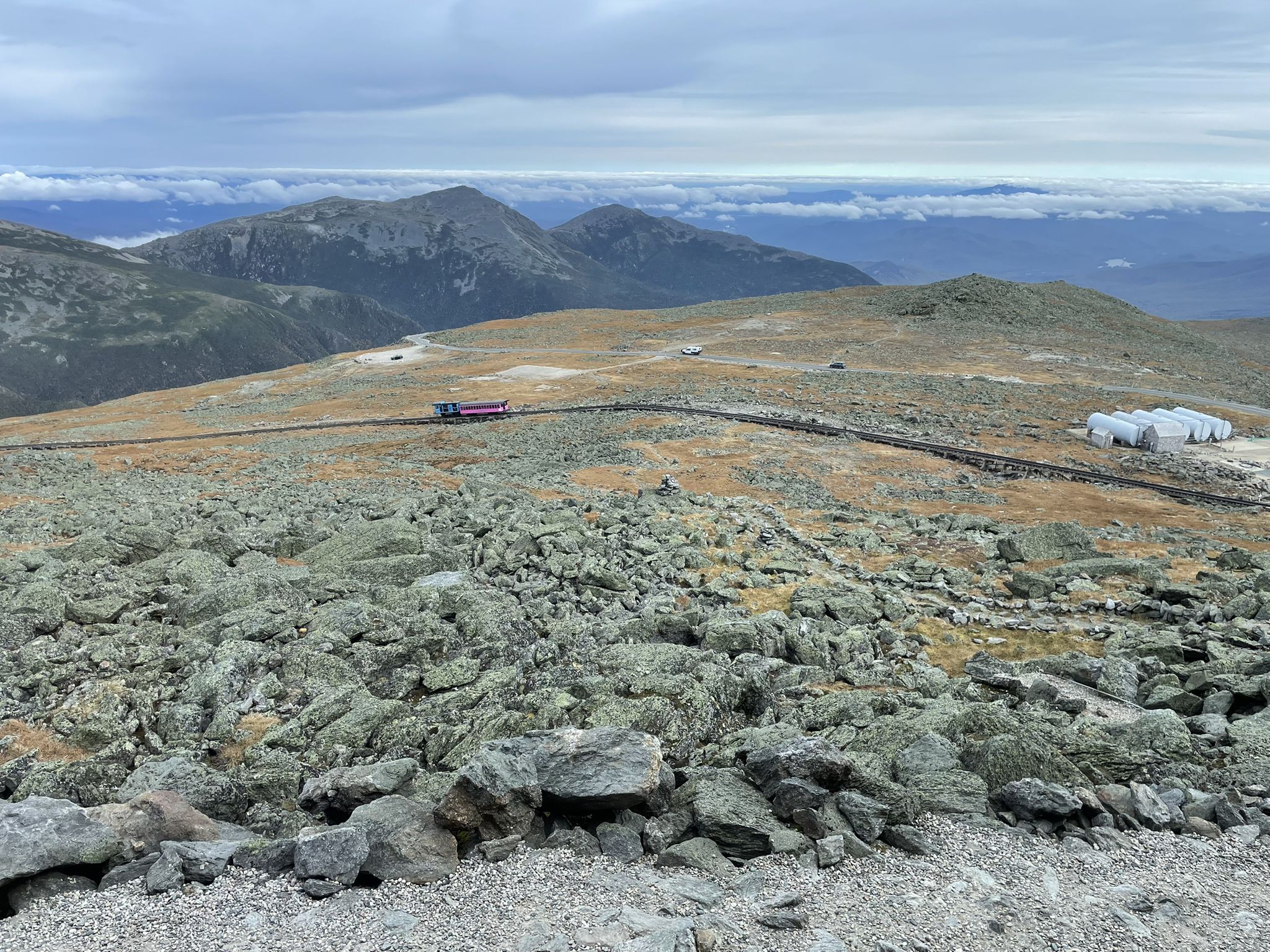 A train on tracks crosses a rocky mountain landscape under a cloudy sky. - Mount Washington via Tuckerman Ravine Trail