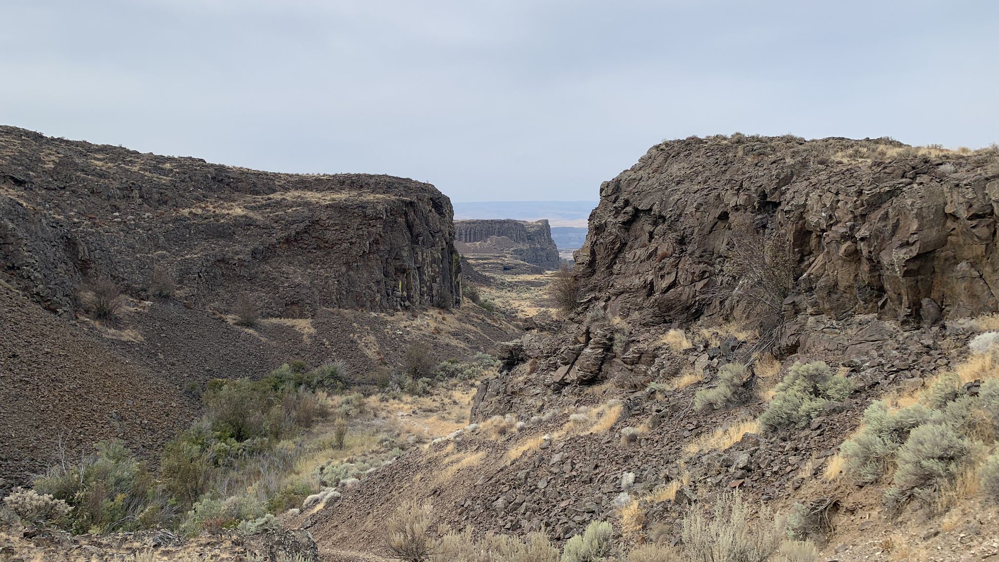 A narrow, rocky canyon with steep walls and dry vegetation under an overcast sky. - Dusty Lake Trail