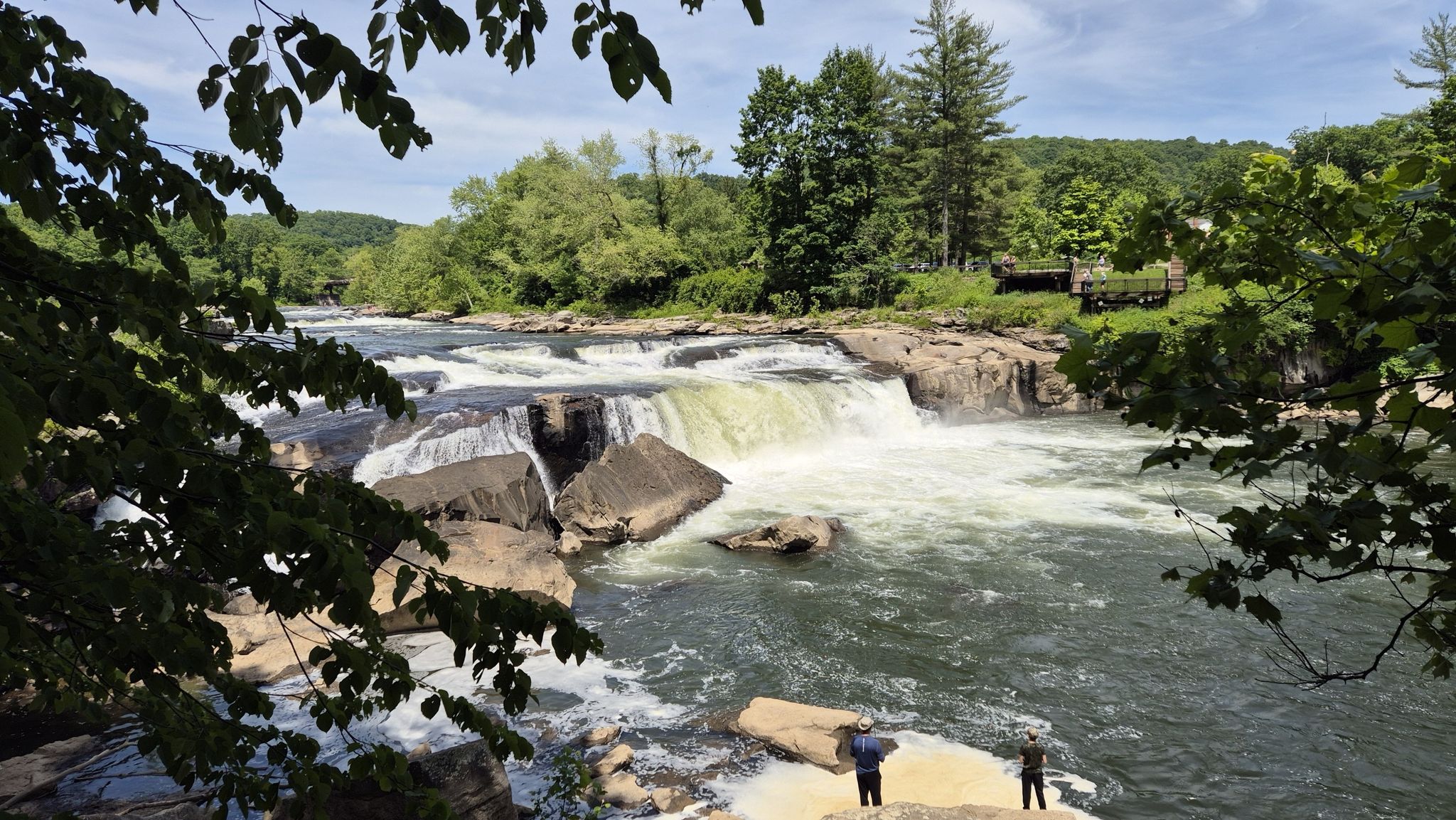 Two people observe a wide waterfall and river under bright daylight. - Ferncliff Trail