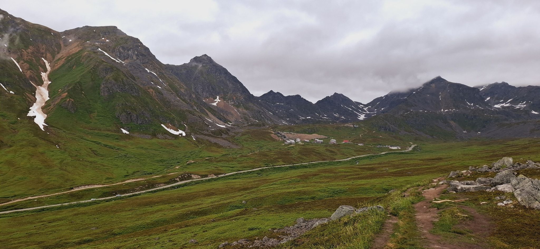 Vast green mountain valley with snow patches, a winding road, under an overcast sky. - Rae-Wallace Mine Trail