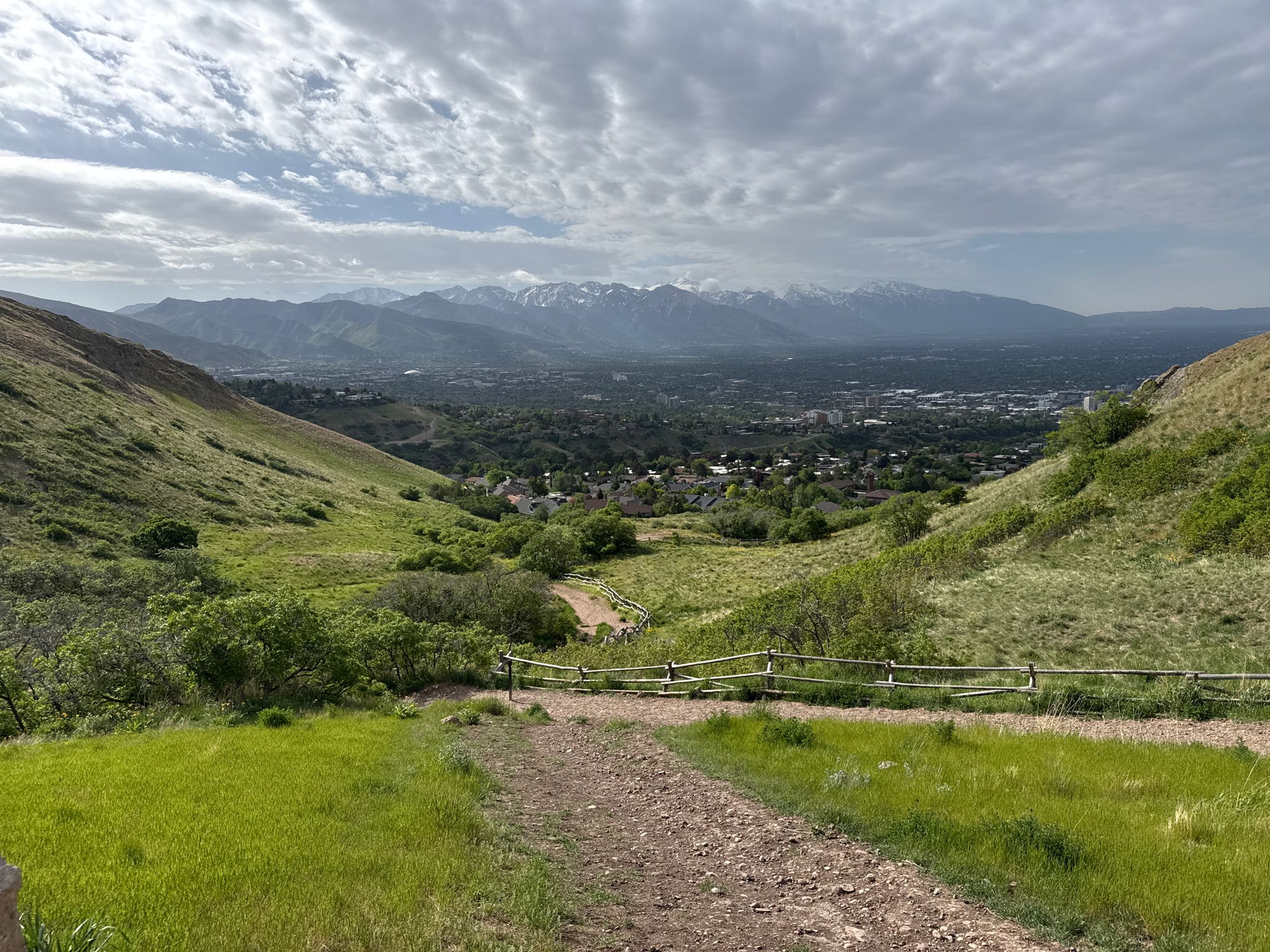 Dirt path descends into a green valley with a city and mountains beyond. - Ensign Peak Trail