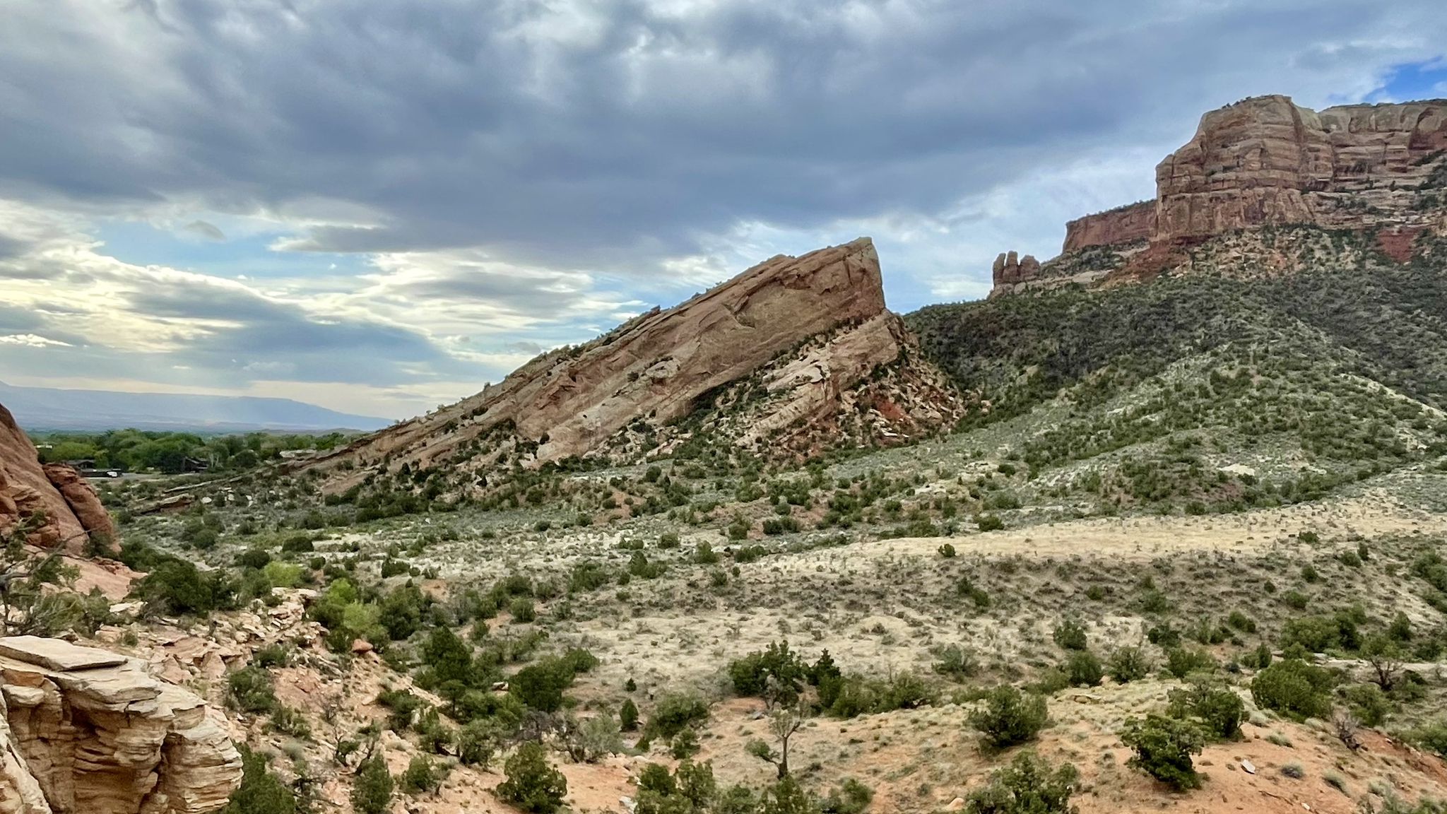 Wide desert vista with sloping red rock formations and green scrub under cloudy skies. - Monument Canyon Trail