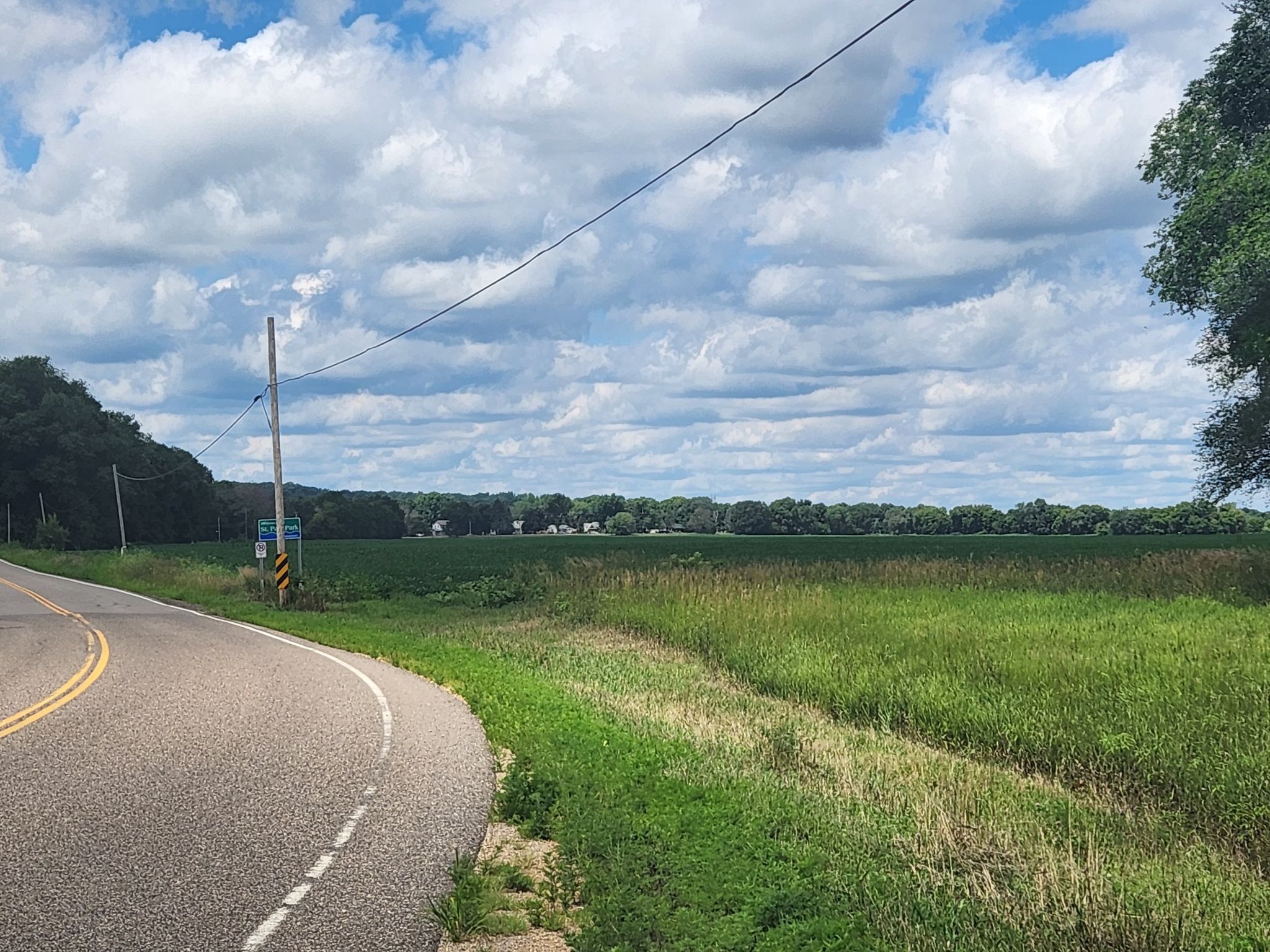 Curved road next to a vast green field under a cloudy sky. - Hazen P. Mooers Park loop from Applewood Neighborhood Preserve