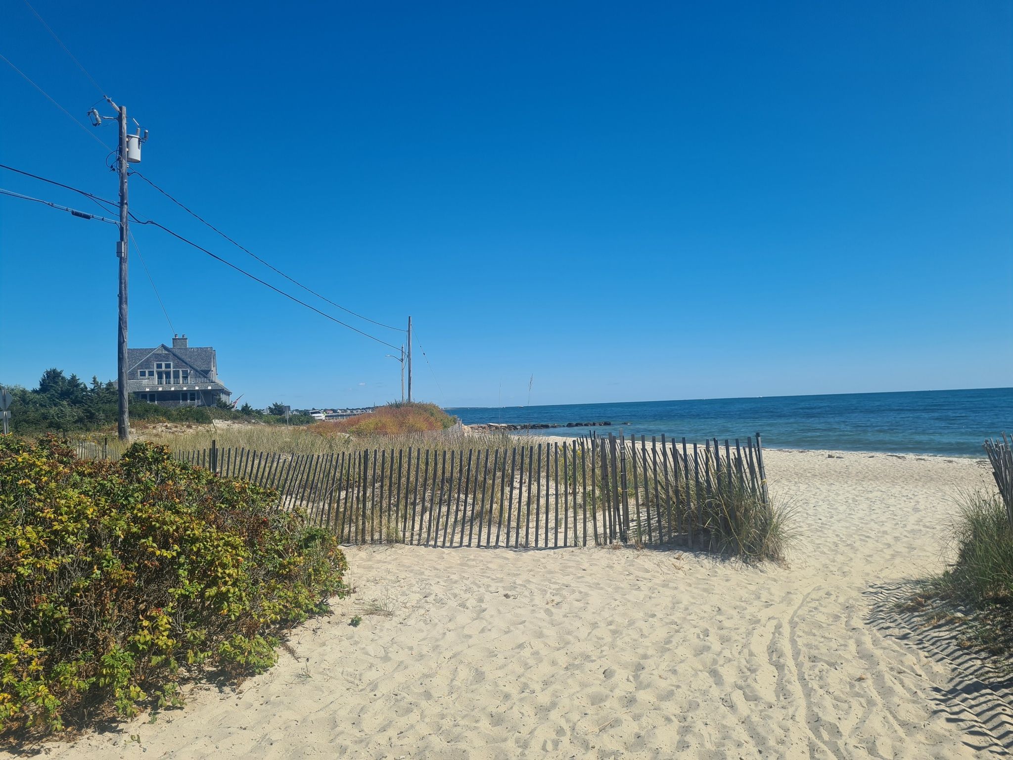 Sandy path with a wooden fence leads to a bright, sunny beach and ocean. - Quissett Beach – Oyster Pond loop from Salt Pond Frontage