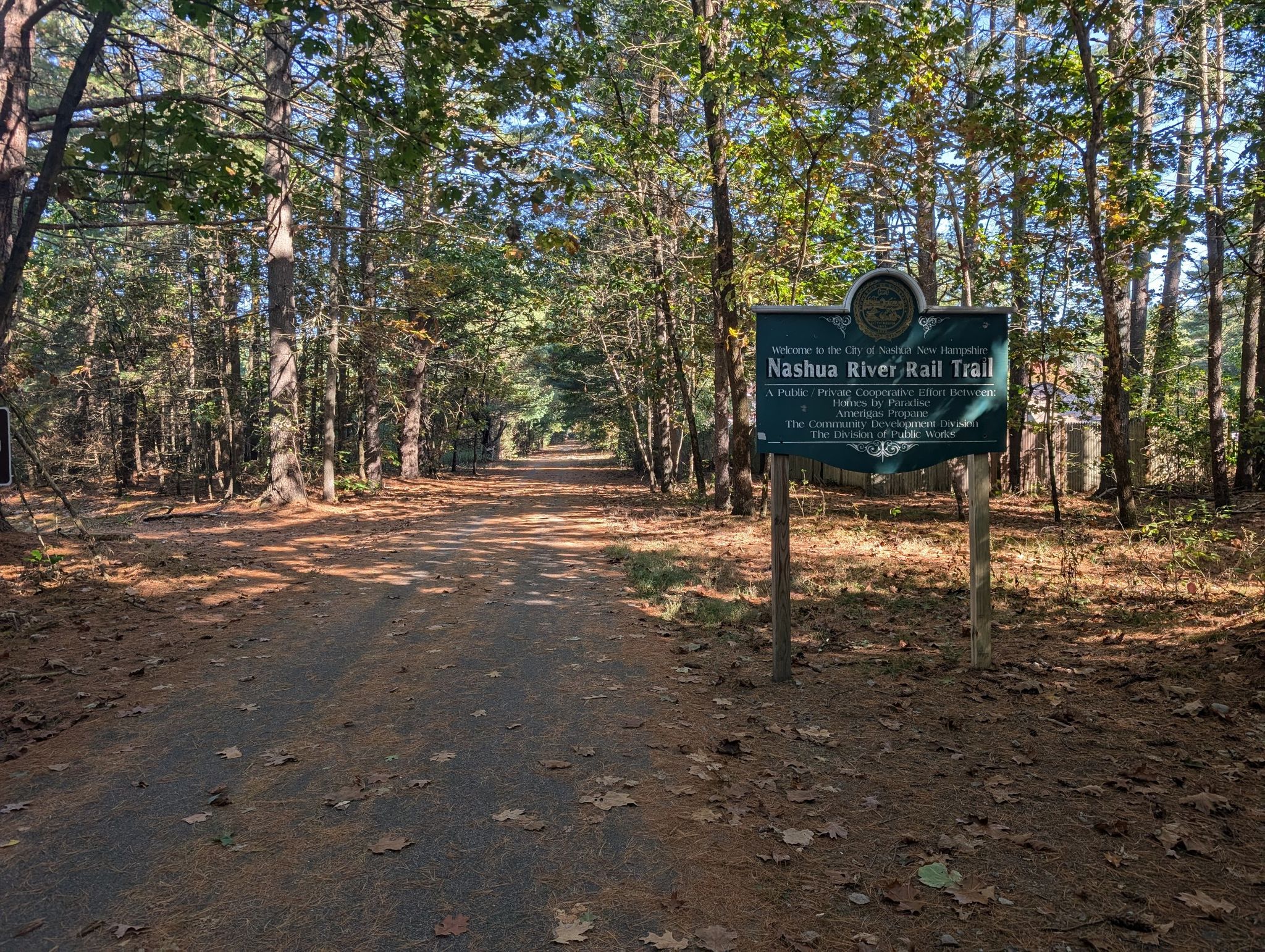 A wide dirt path through a forest with a trail sign on the right. - Nashua River Rail Trail