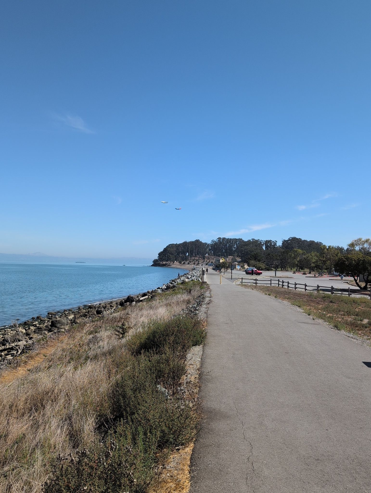 Paved path along a rocky shore, leading towards a tree-covered point. - Promenade Trail and Bluff Loop