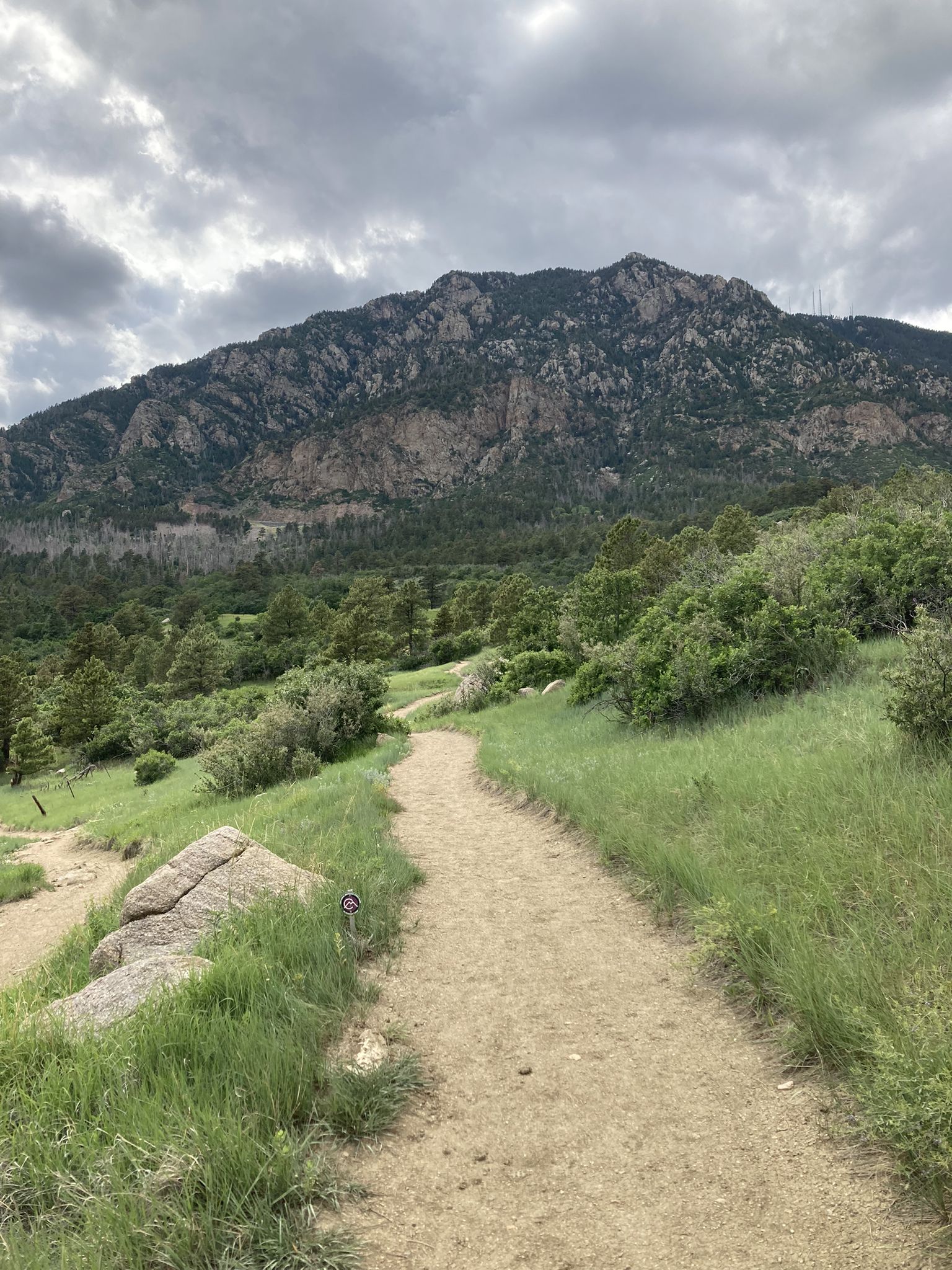 A dirt path winds through green grass towards rocky mountains under a cloudy sky. - Cheyenne Mountain State Park loop