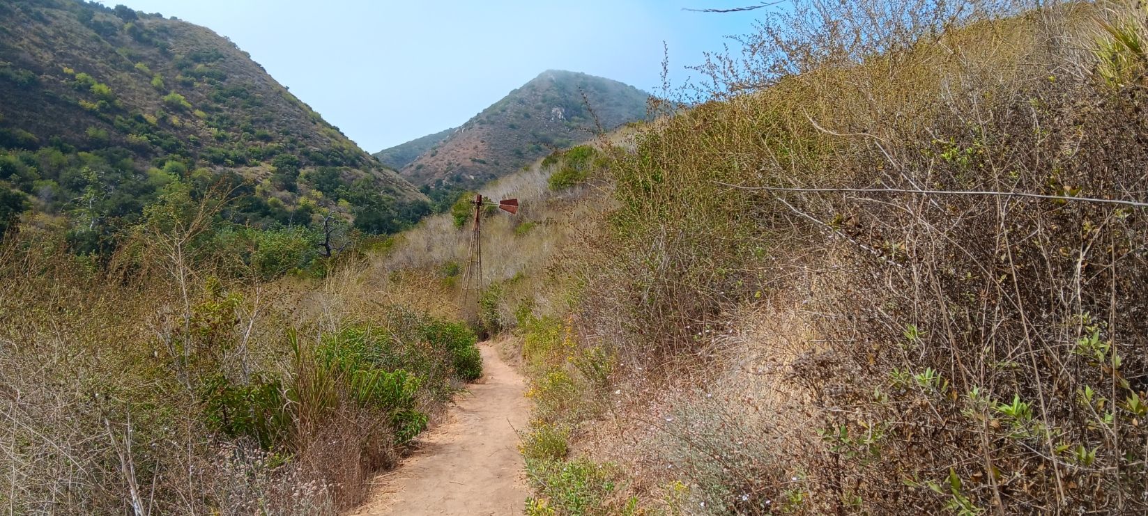 Dirt path through dry brush leading into a valley with hazy hills. - TRW Loop Trail