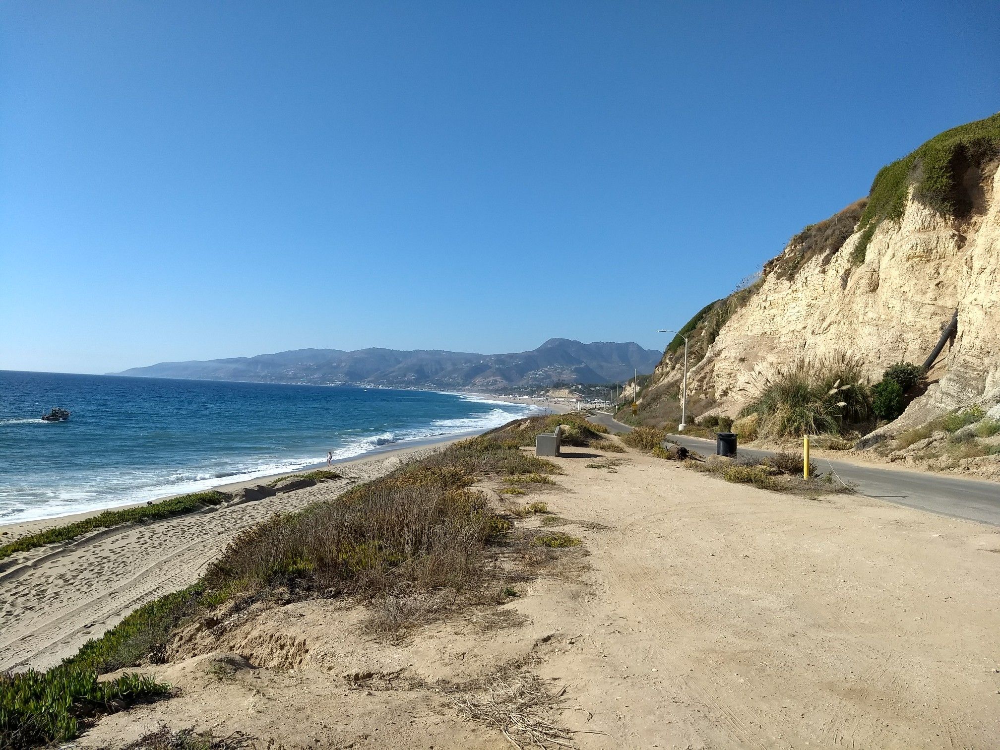 Sunny beach with ocean waves, distant mountains, a boat, and a road by a cliff. - Westward Beach