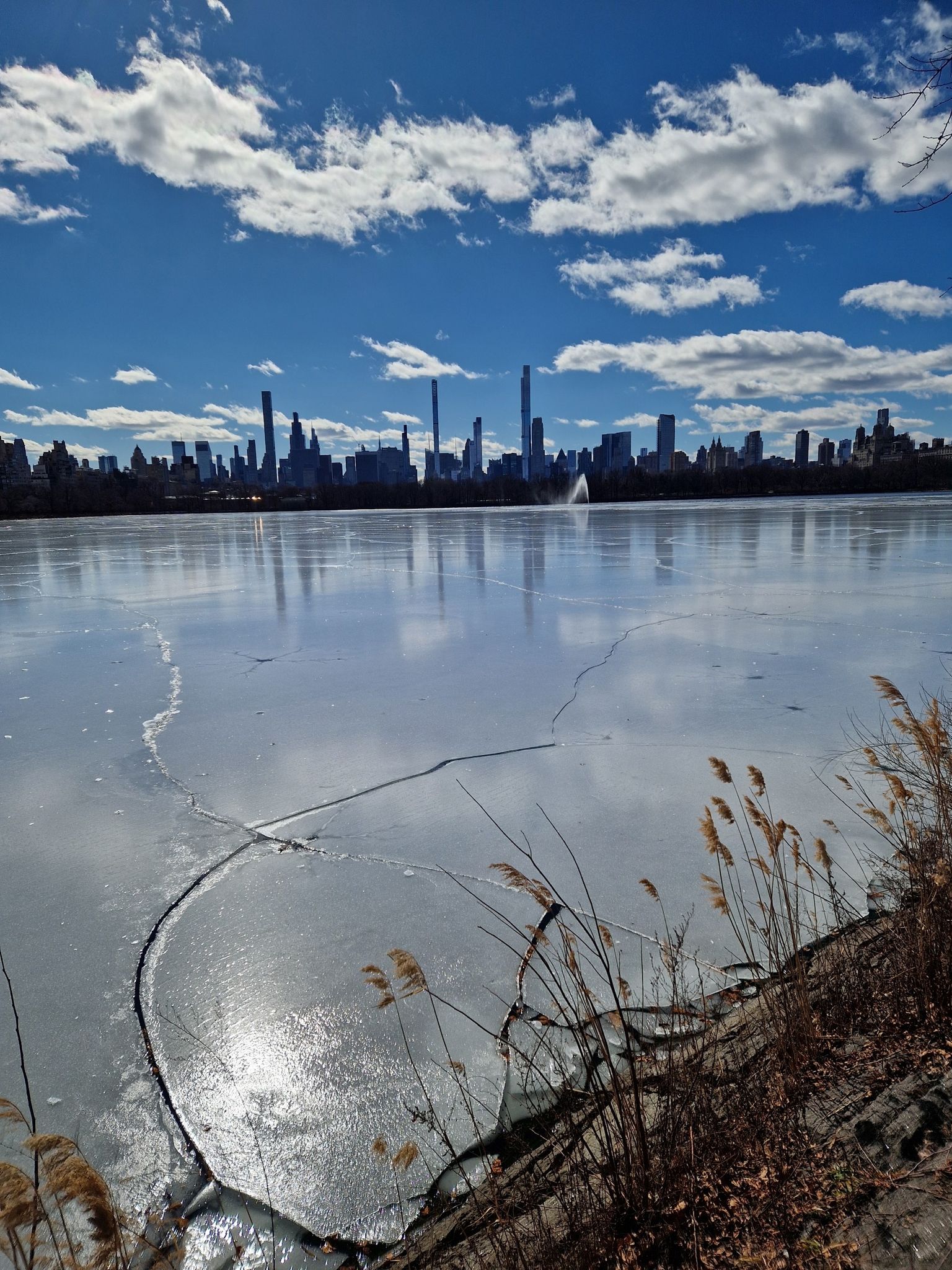 Partially frozen lake with a distant city skyline under a bright blue sky. - The Pool, Central Park – Harlem Meer loop from Cathedral Parkway–110th Street
