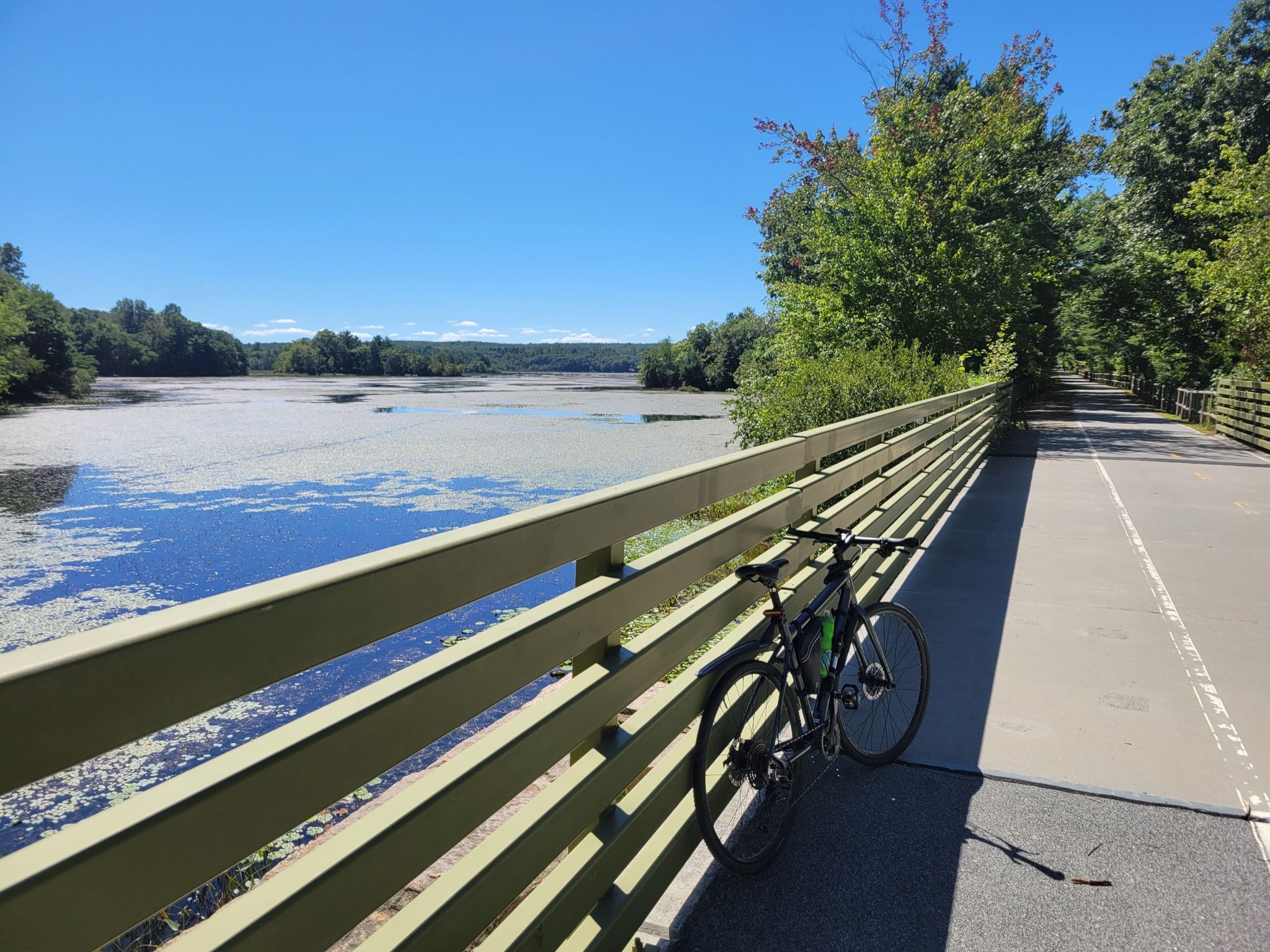 Bicycle resting on a bridge railing overlooking a wide, sunny, algae-covered river. - Trestle Trail loop from Coventry