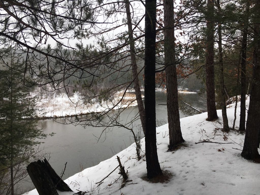 Overcast winter scene of a river flowing past snow-covered banks and pine trees. - Fife Lake Loop