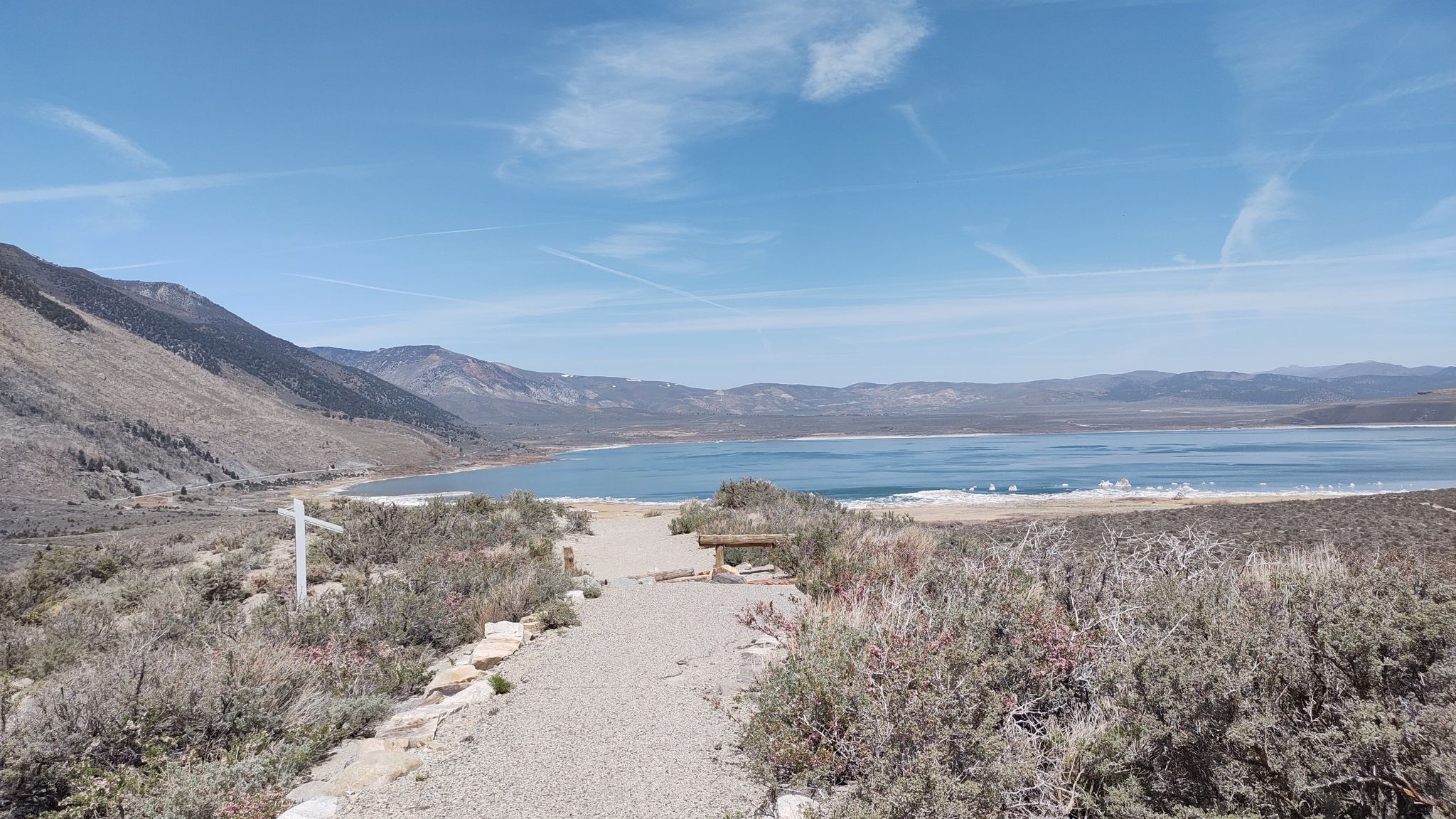 A gravel path leads to a large lake surrounded by arid mountains under a blue sky. - Mono Lake Trail