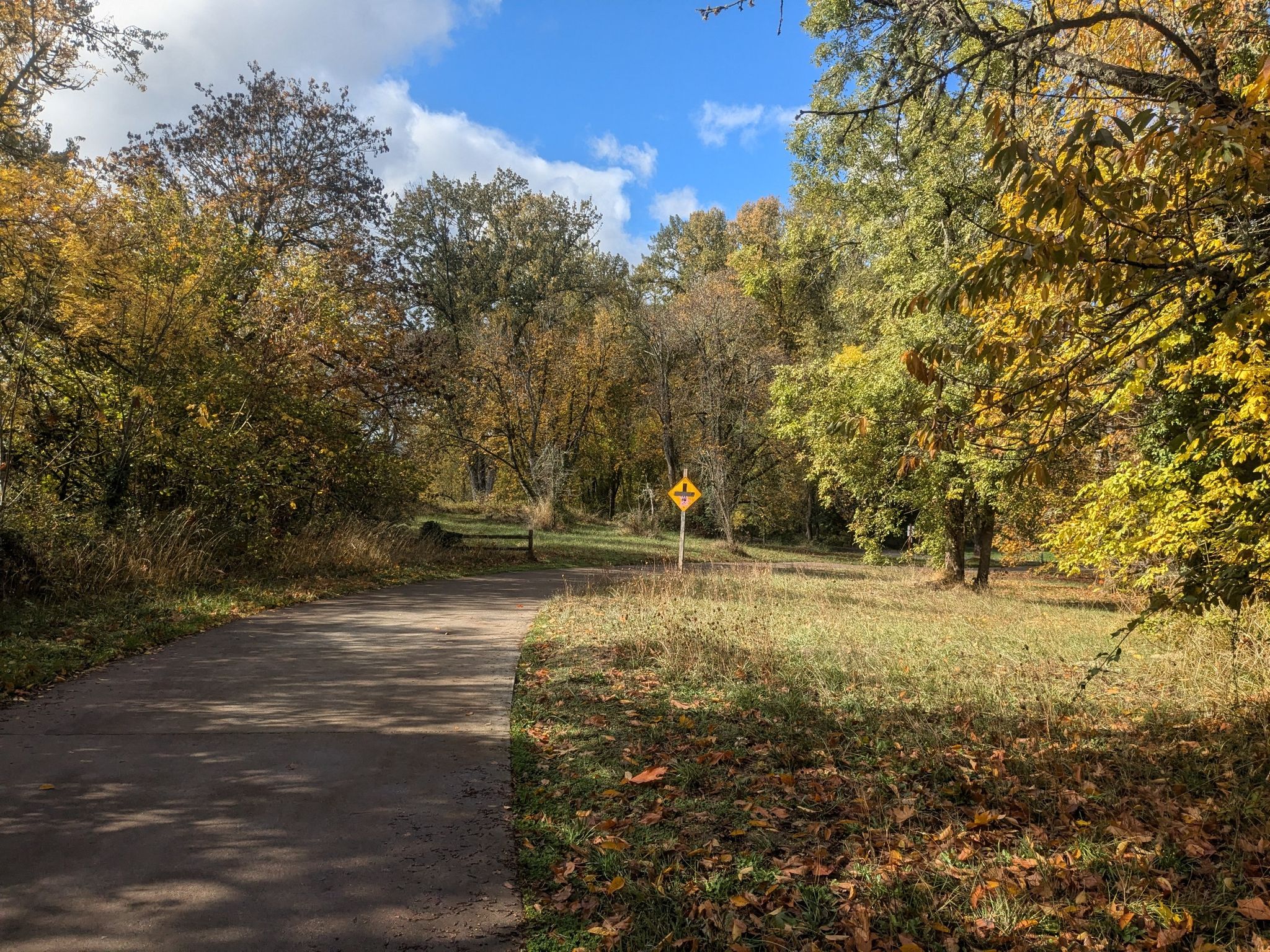 Paved path through autumn woods with a yellow warning sign. - Frohnmayer Bridge – Alton Baker Park loop from Gateway Mall