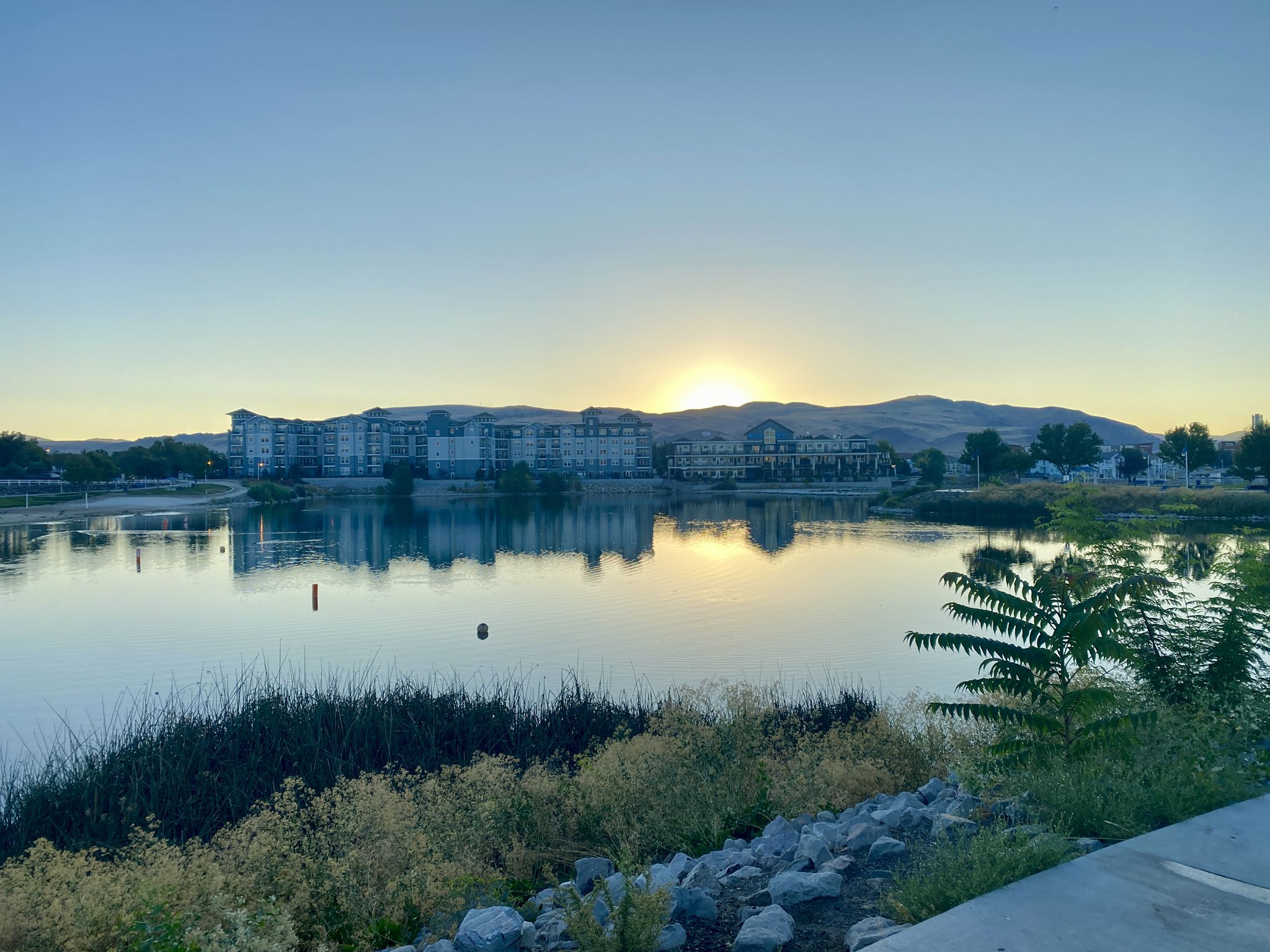 Sunrise over a calm lake with distant buildings and mountains. - Sparks Marina Loop Trail