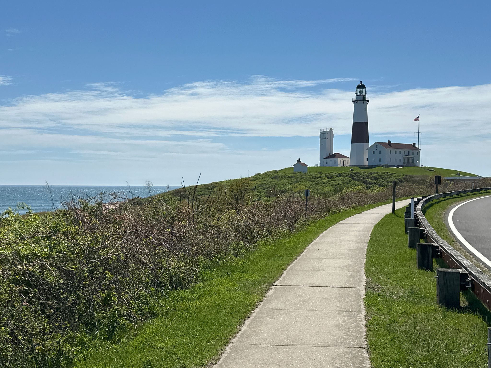 Lighthouse and buildings on a grassy hill by the ocean on a bright day. - Montauk Point Lighthouse – Hither Hills State Park loop from Montauk