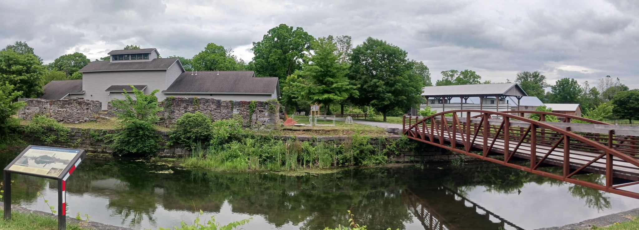 A canal with buildings, a metal bridge, and green trees under an overcast sky. - Erie Canal – Canal Overpass loop from Canastota Reservoir