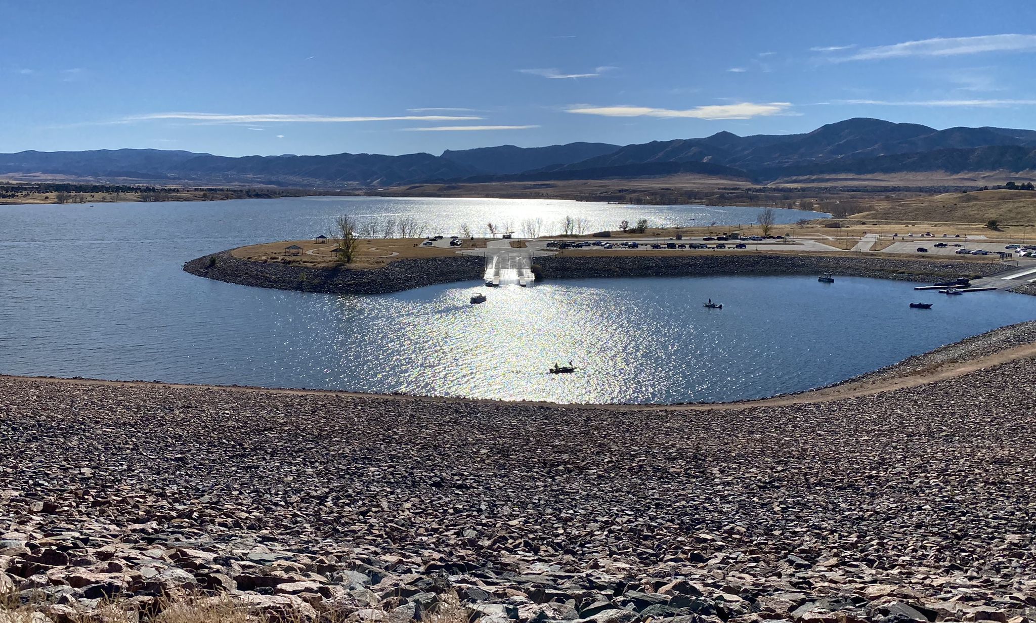 Sunny lake with boats, rocky foreground, and distant mountains under bright sky. - Chatfield Reservoir loop