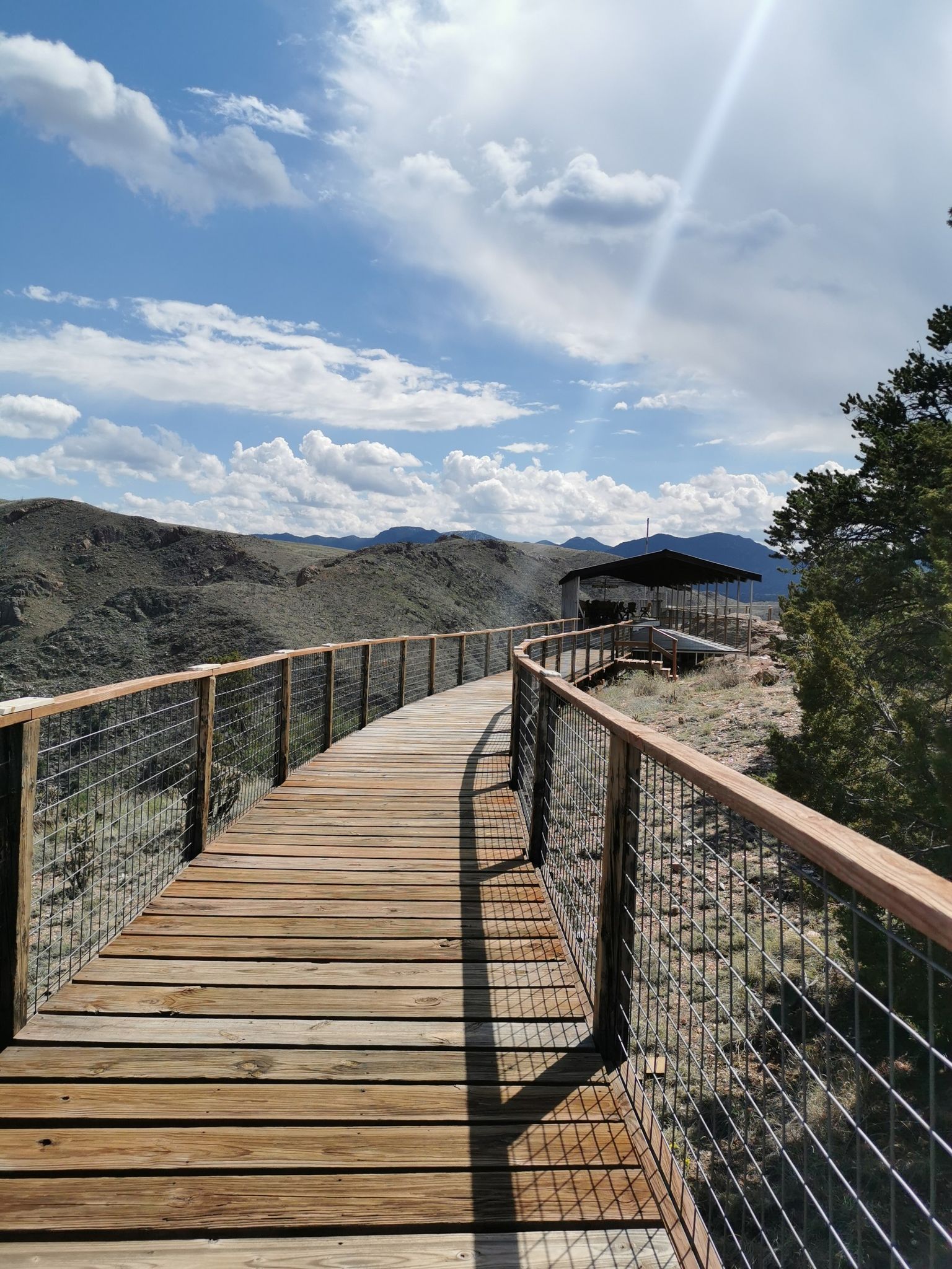 Curved wooden walkway leading to a covered structure on a sunny, hilly landscape. - Point Alta Vista Trail loop from Buckskin Joe