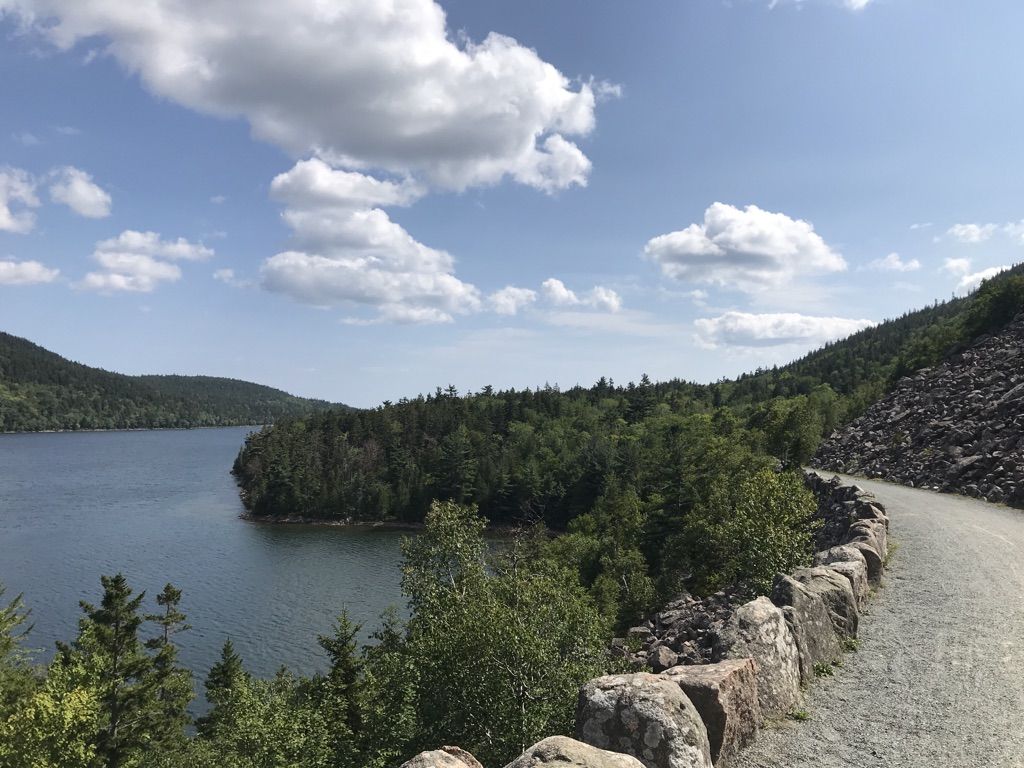 Gravel path and stone wall beside a calm lake and green forest. - Tri Lakes Loop