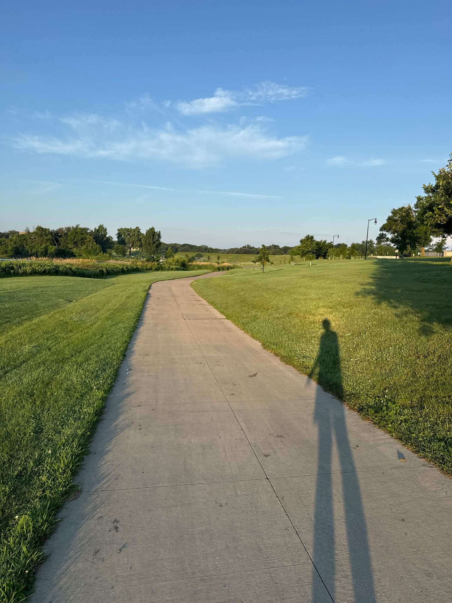 Paved path through green fields under a clear sky, with a long human shadow. - Red River Trail Loop