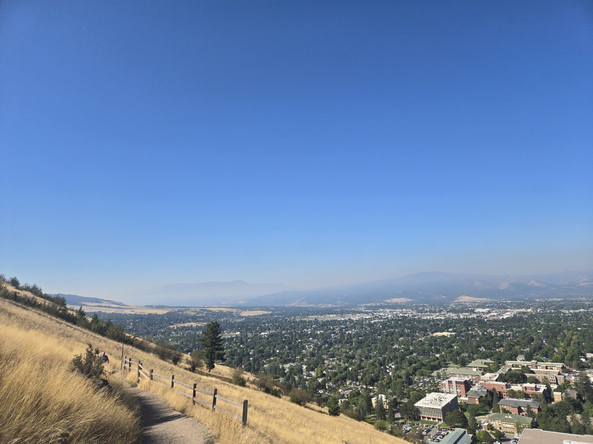A sunny, dry hillside path overlooks a green valley town and hazy mountains. - The M Trail on Mount Sentinel loop from Missoula