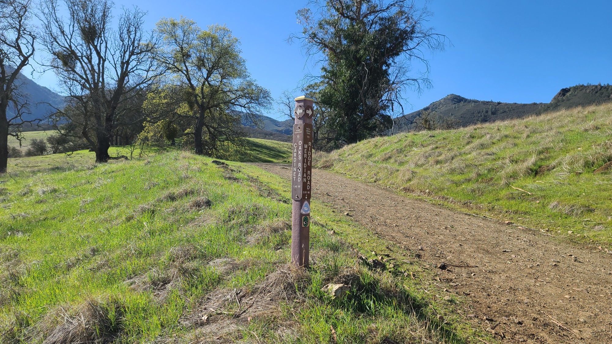 A dirt path next to a wooden post in a sunny green field. - Clayton Oaks, Donner Canyon, and Bruce Lee Spring Loop