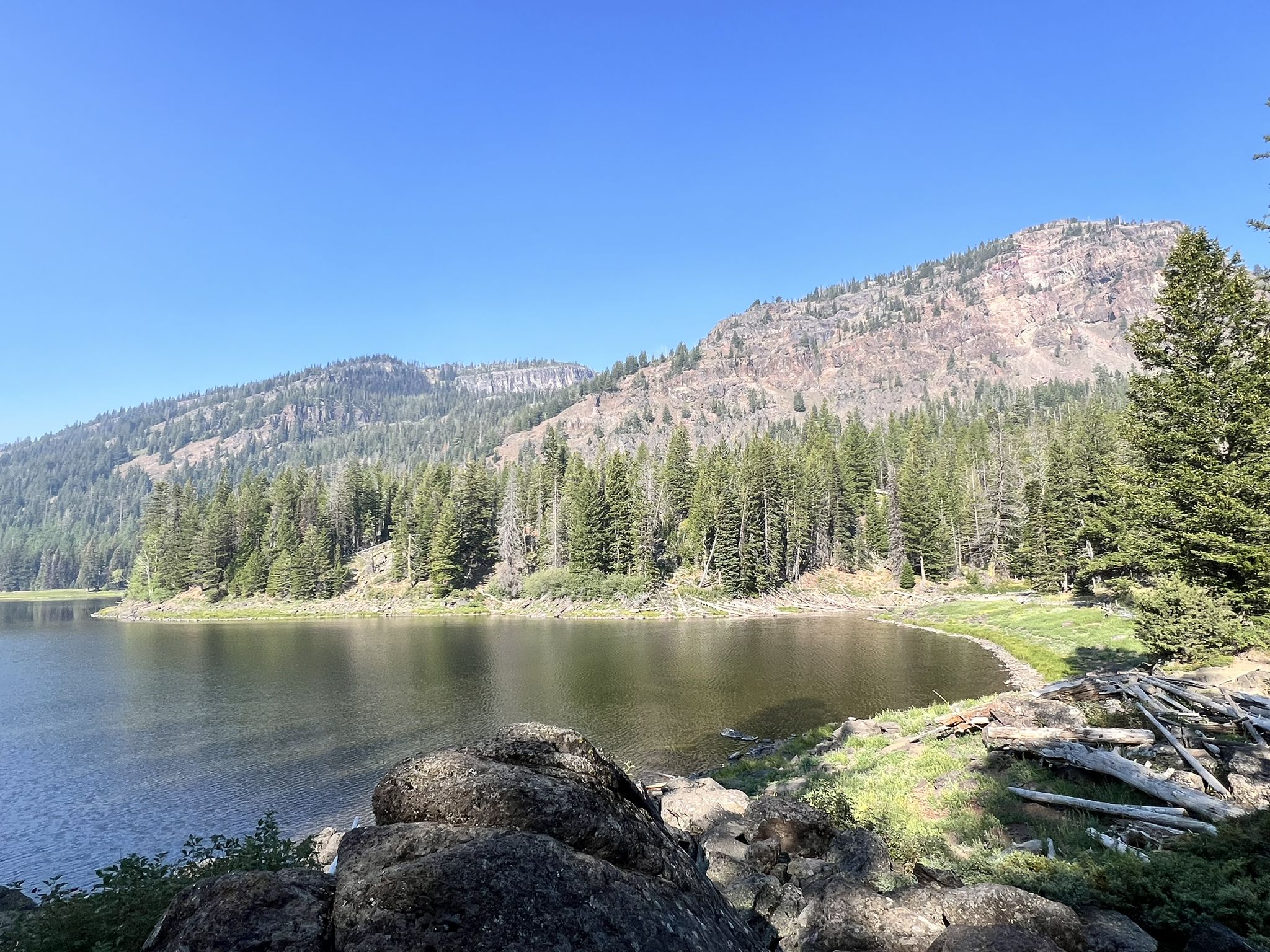 Serene lake with rocky foreground, forested mountains, and clear blue sky. - Slide Basin Trail