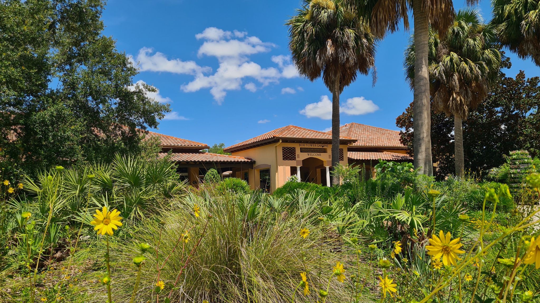 Building with red tile roof behind tropical plants and yellow flowers, sunny. - Bok Tower Loop