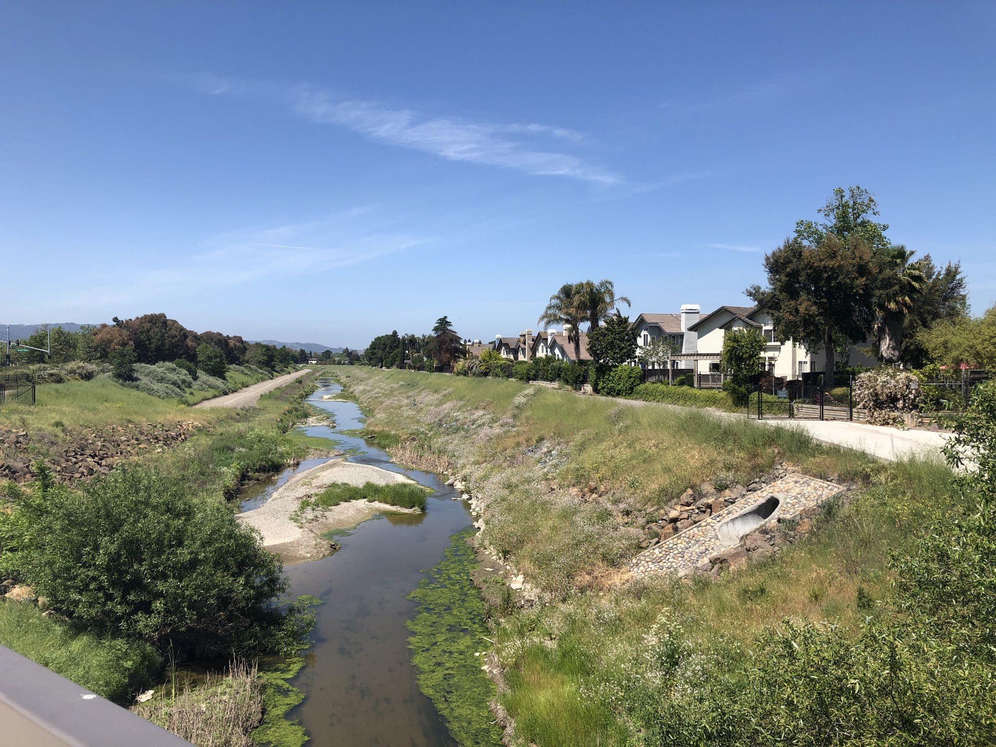 A narrow river flows between grassy banks with houses under a clear sky. - Iron Horse Regional Trail