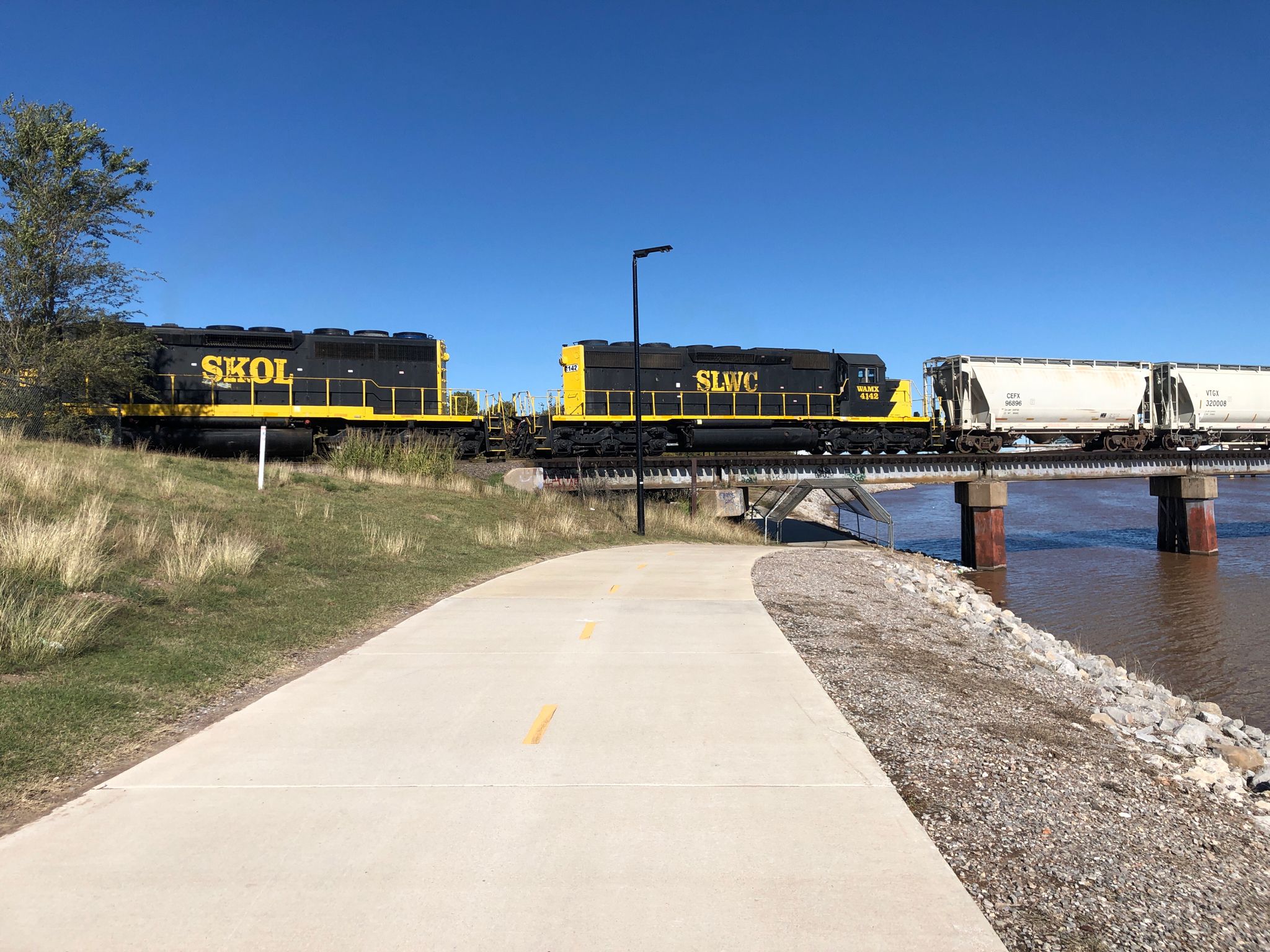 Train on a bridge over a river next to a paved path. - Oklahoma Land Run Monument – Regatta Park loop from Oklahoma City