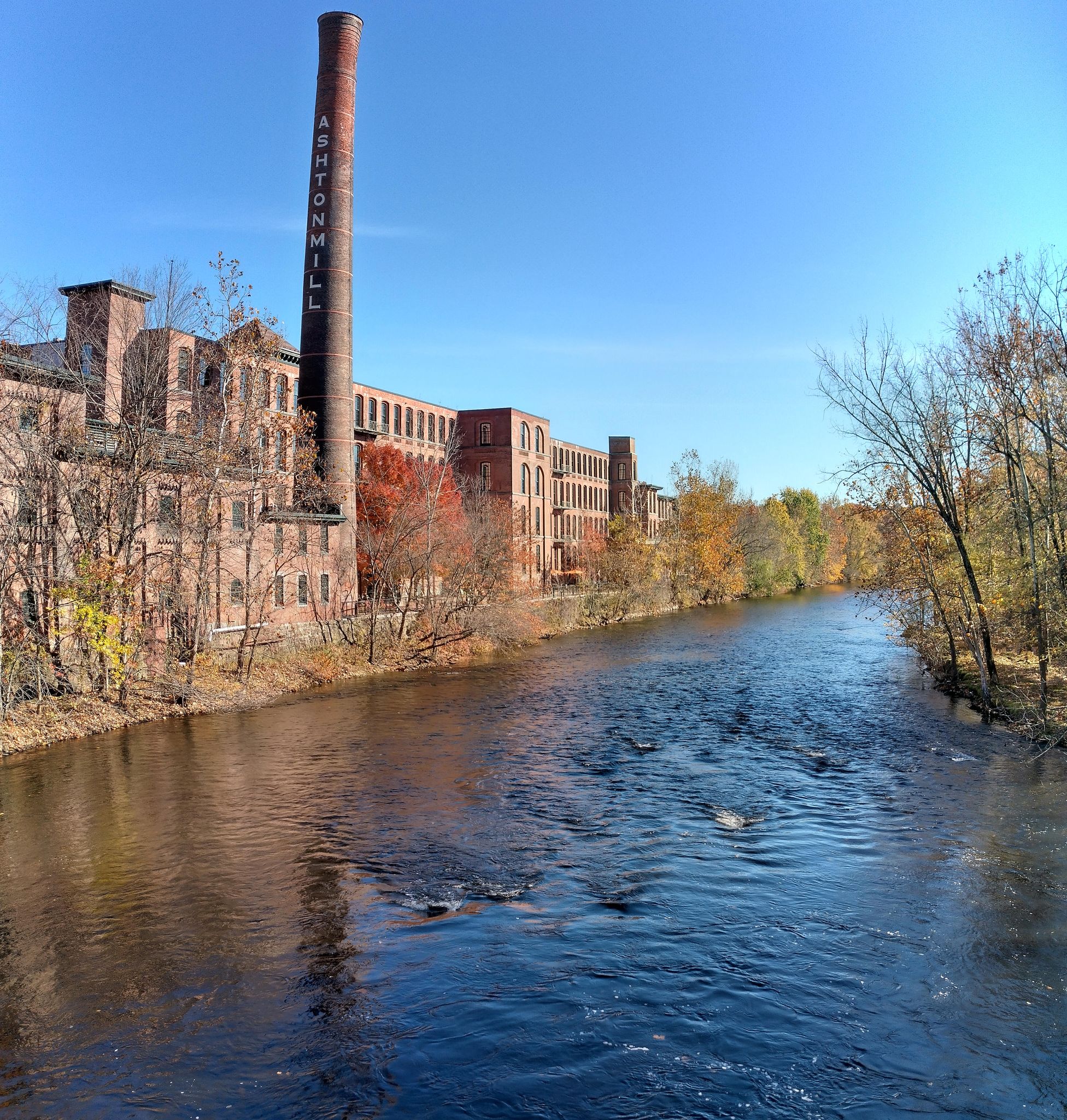 An old brick mill building and smokestack stand beside a river with autumn trees. - Along the Blackstone River – Blackstone River Bikeway loop from Albion